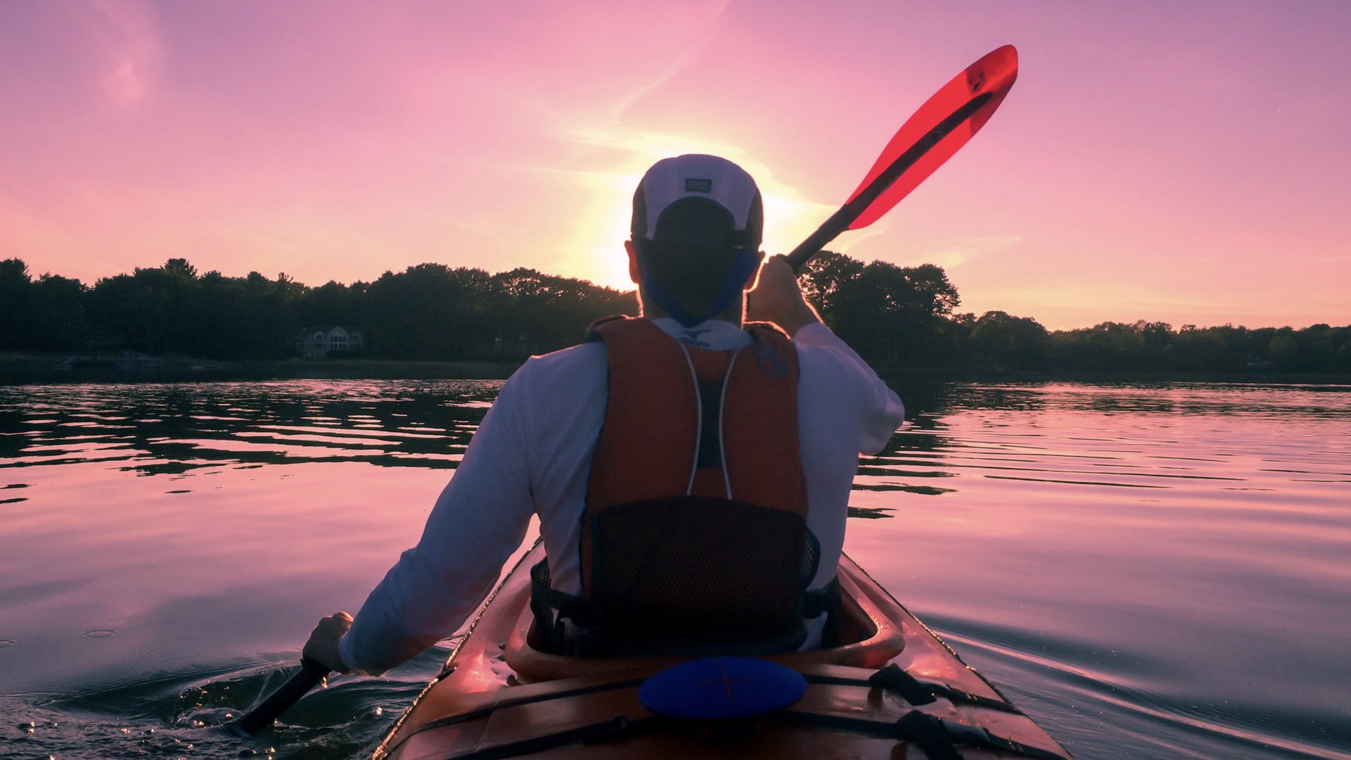 Person kayaking on a body of water during sunset or sunrise, holding a paddle, with trees in the background and the sky painted in pink and orange hues.