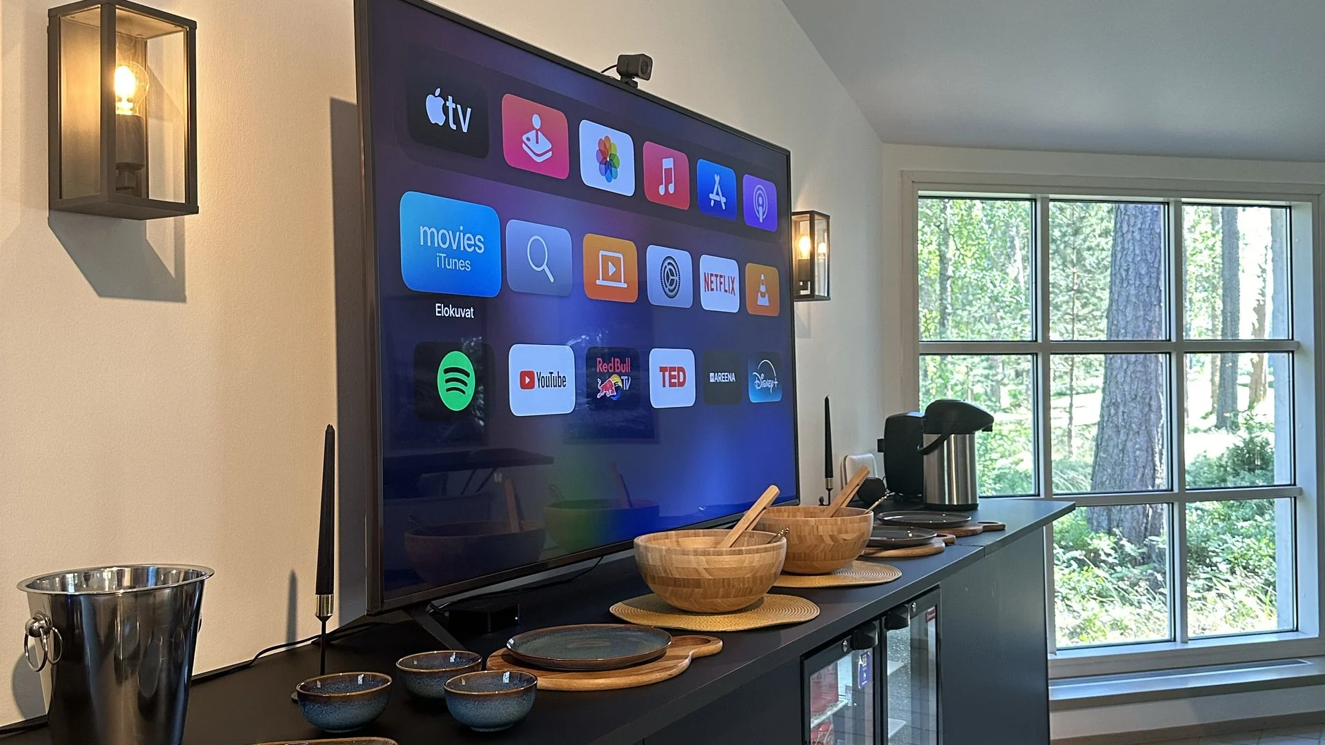 Living room with a flat-screen TV displaying various streaming app icons, next to a window with trees outside, and decorated with wall sconces, bowls, and kitchen appliances.