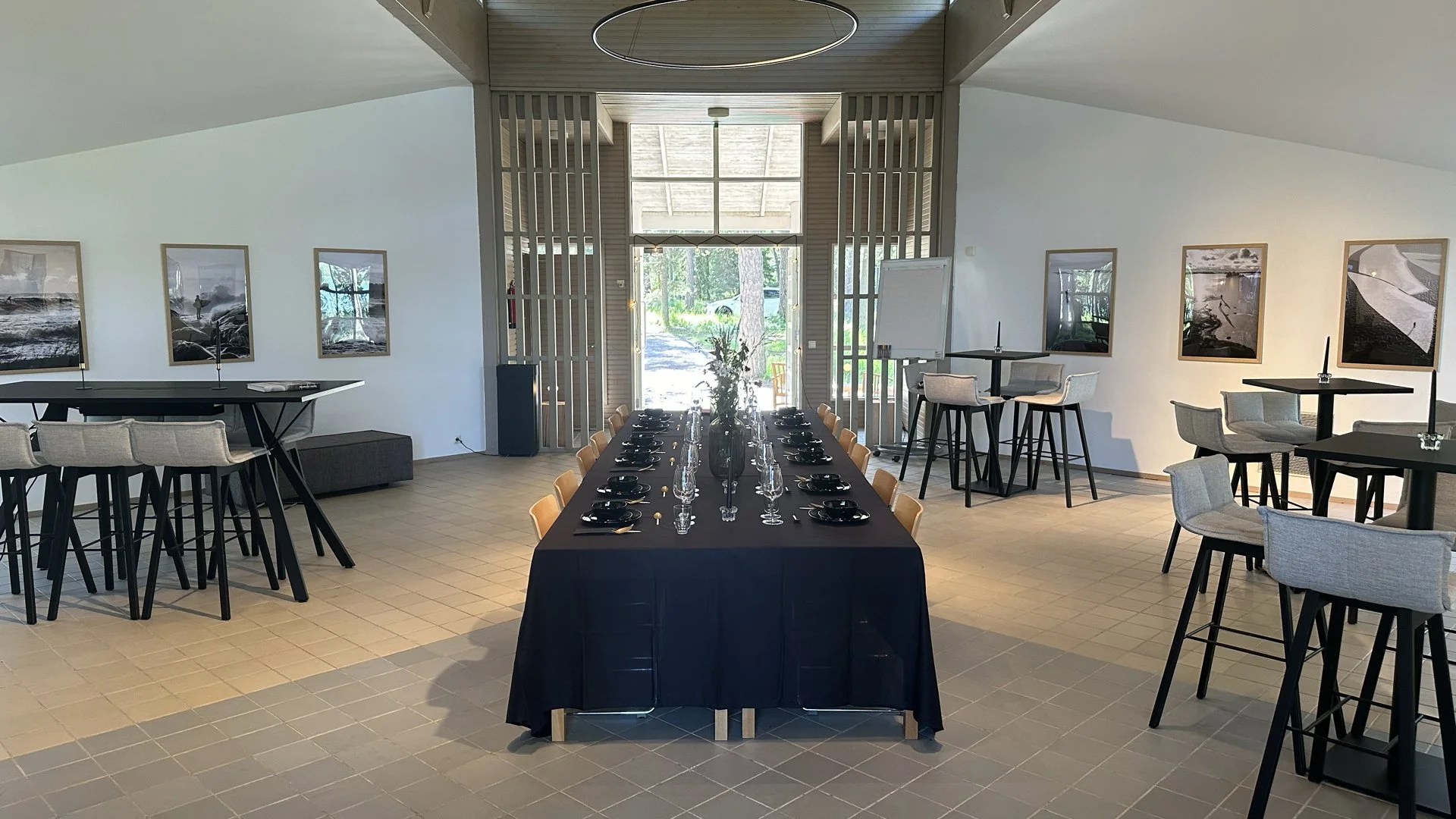 Event space with a long table set for dining, surrounded by chairs, and high tables with bar stools, large windows, and framed artwork on white walls.