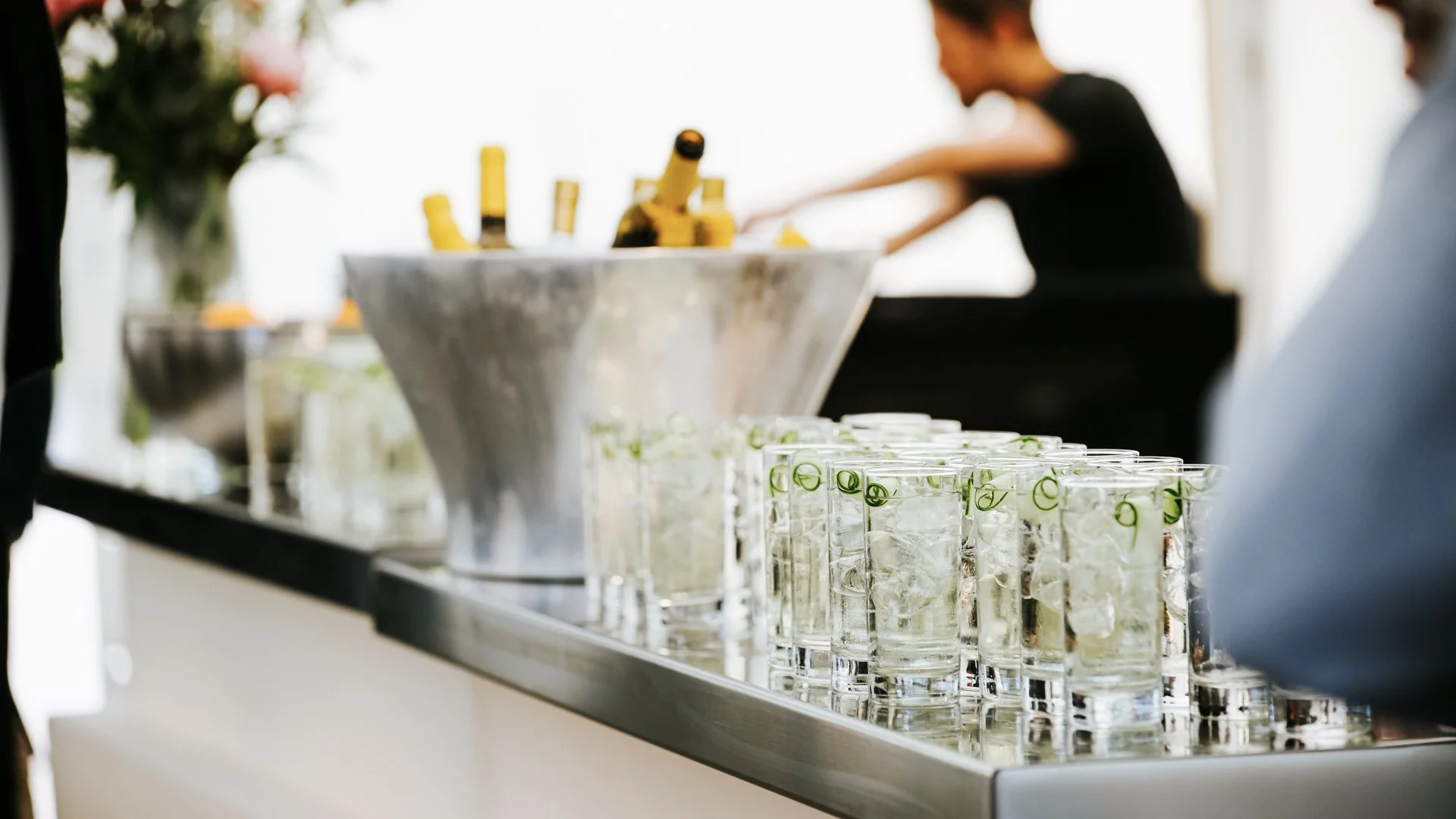 A row of shot glasses filled with ice and cucumber slices on a bar counter, with blurred background including a person preparing drinks and bottles.