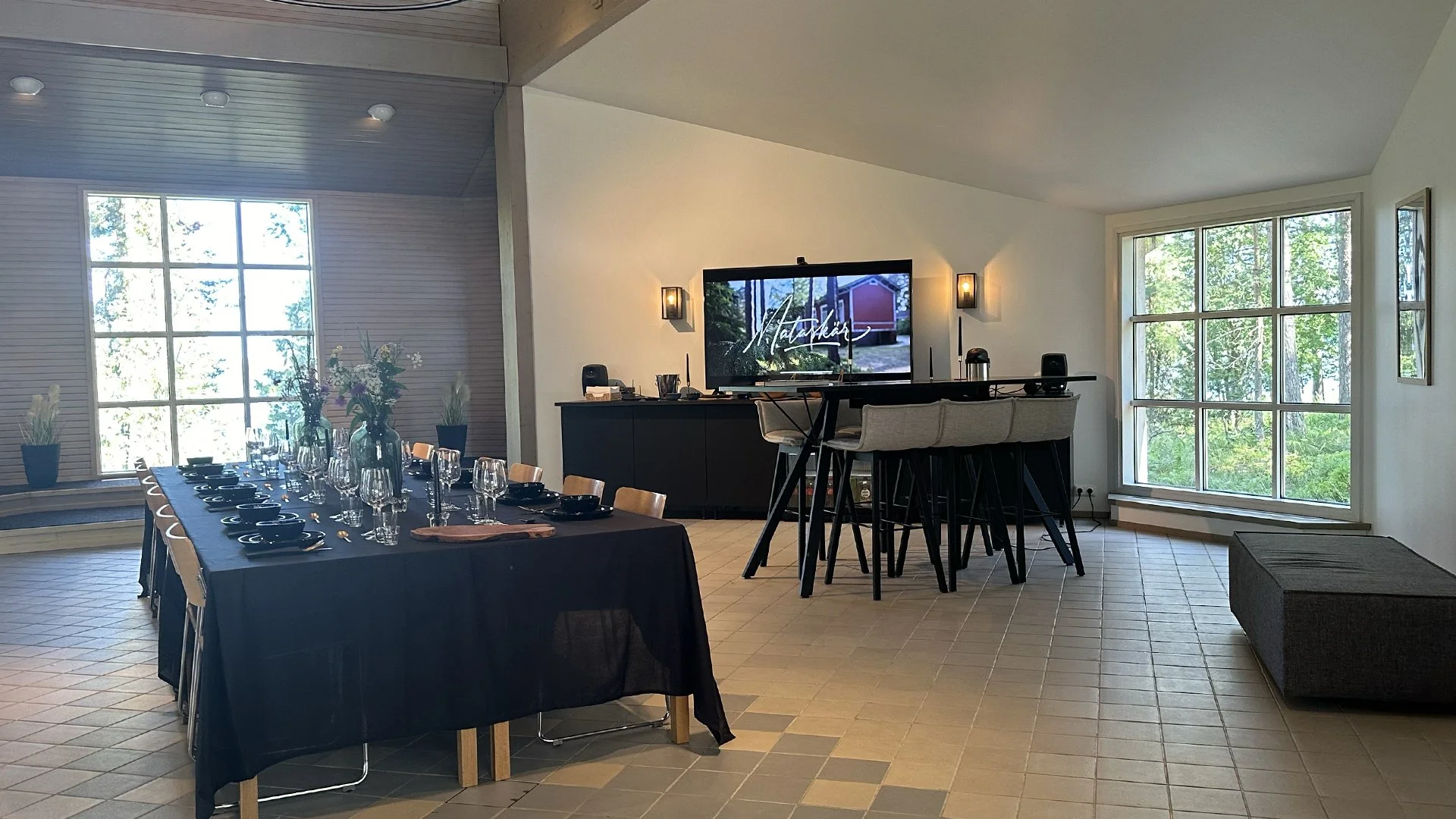 A dining and entertainment area set up for a gathering with a long table covered with a black tablecloth, black plates, glasses, and floral centerpieces. A large window with trees outside, a television on a black cabinet, and a high table with bar st