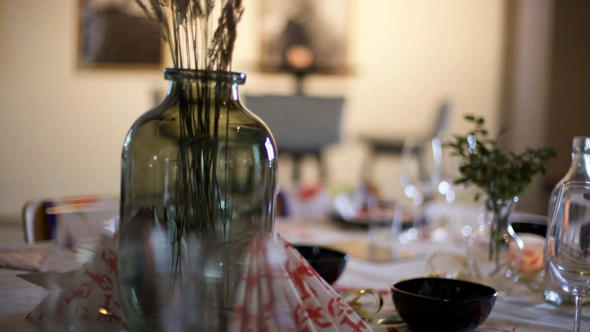 Close-up of a glass vase with dried flowers on a dining table, with blurred table settings and chairs in the background.