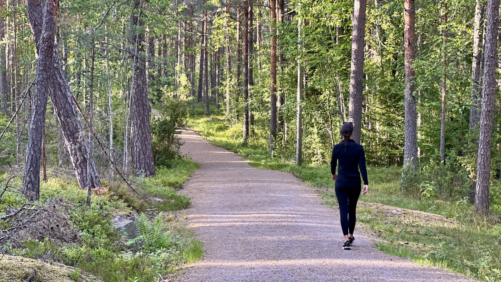 A woman walking on a dirt trail through a dense forest with tall trees and green foliage.