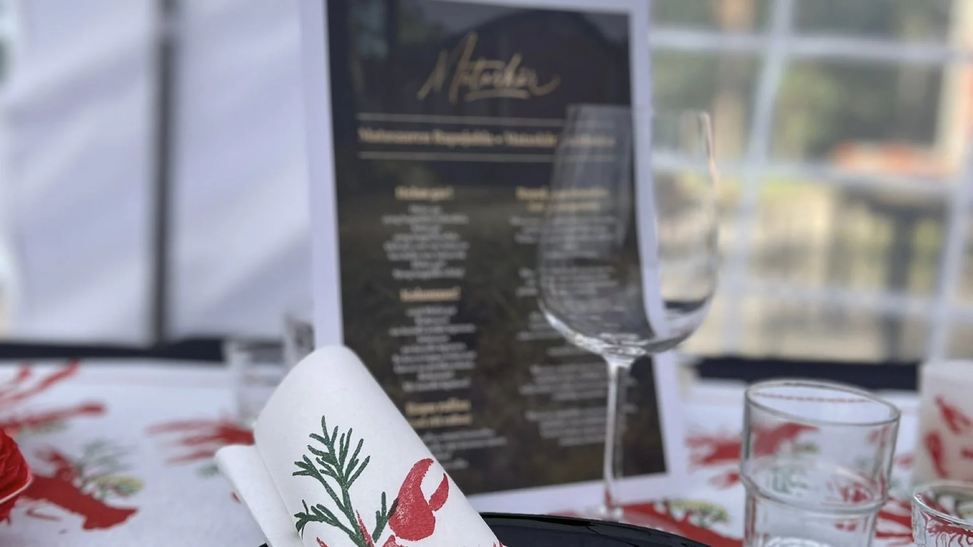 A table set with wine glasses, a menu, and holiday-themed napkins with a red lobster and green herbs design, in a well-lit space with large windows.