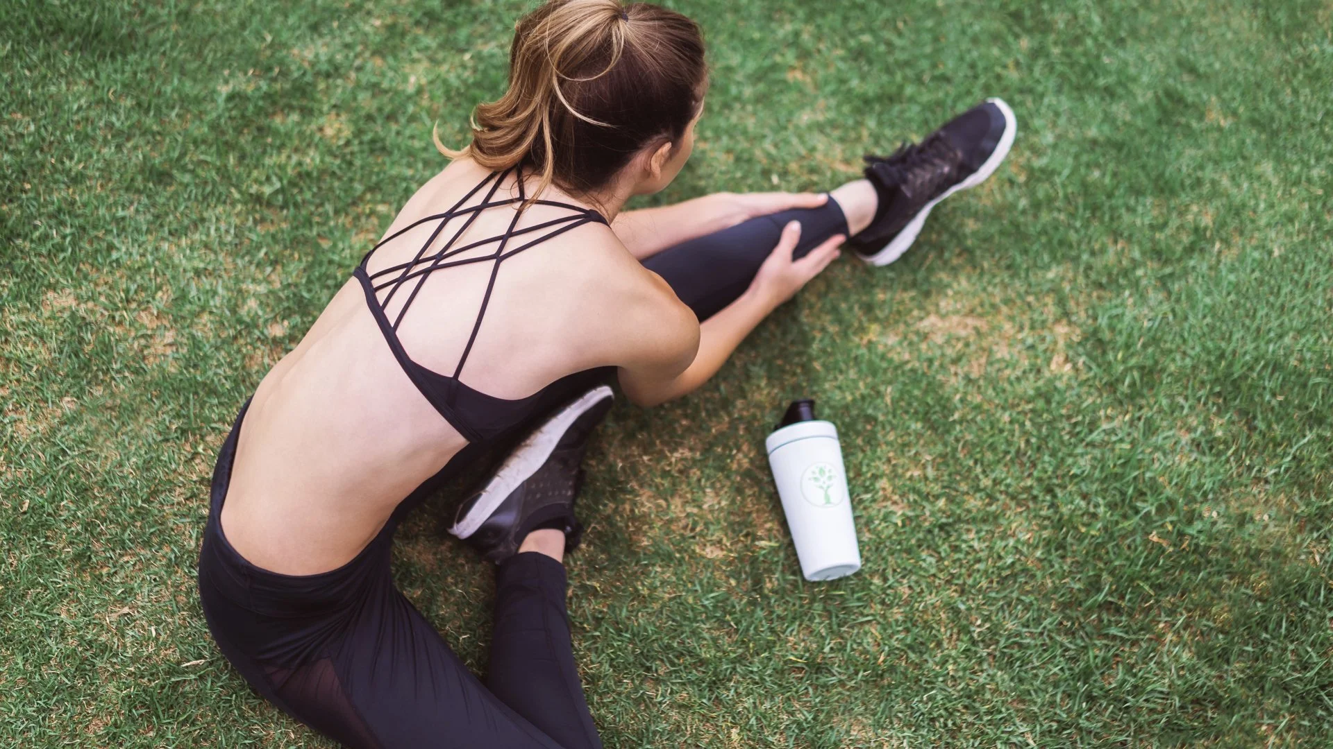 A woman in black athletic wear sitting on grass, stretching with her right leg extended and her left knee bent, near a white water bottle with a green tree logo.