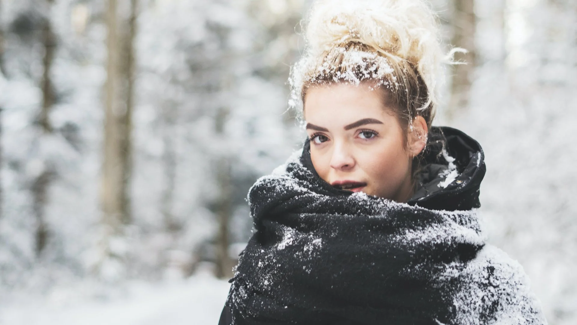 Young woman with blonde hair styled in a messy bun, wearing a black winter jacket, standing in a snowy forest, facing slightly to the right, with snow on her hair and jacket.