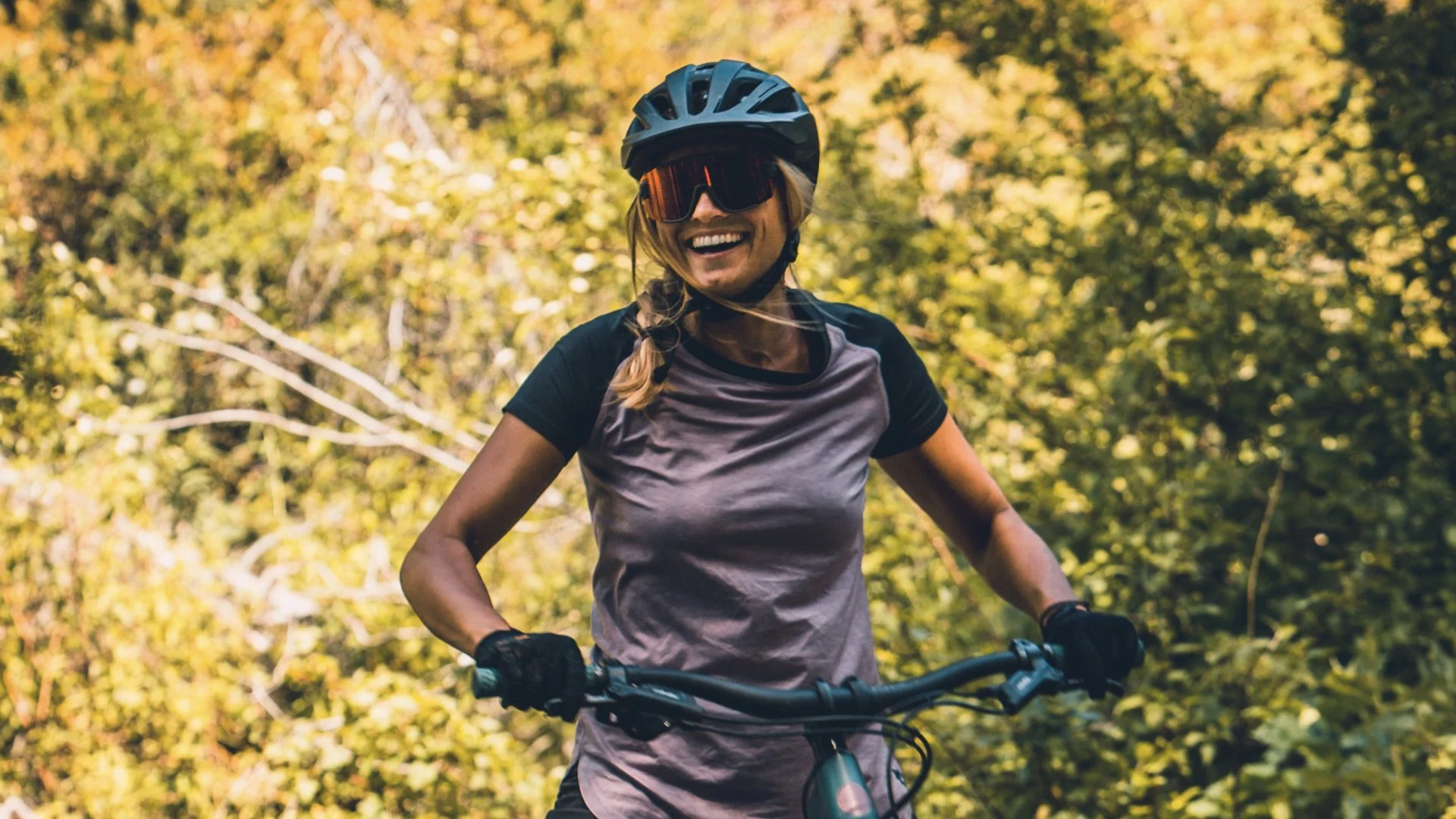A smiling woman wearing a helmet and sunglasses, riding a mountain bike through a forest with green trees and yellow leaves.
