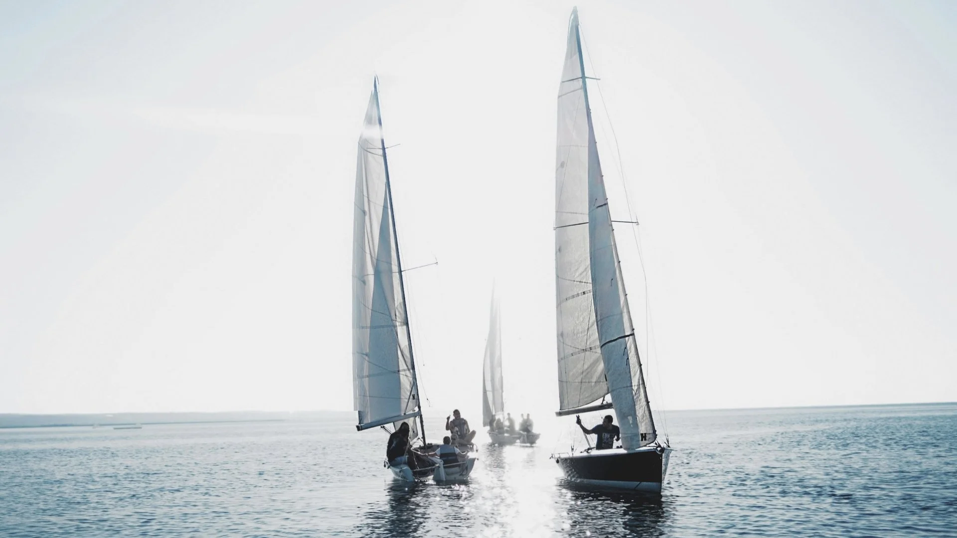 Sailing boats on calm water with a bright sun creating glare, with people on the boats and a distant horizon.