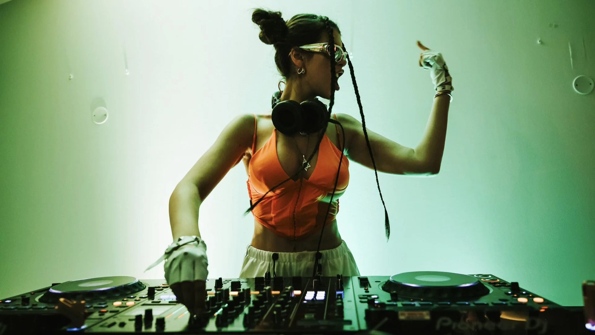 A woman DJing at a club, wearing sunglasses, a bright orange crop top, and white gloves, with colorful lighting