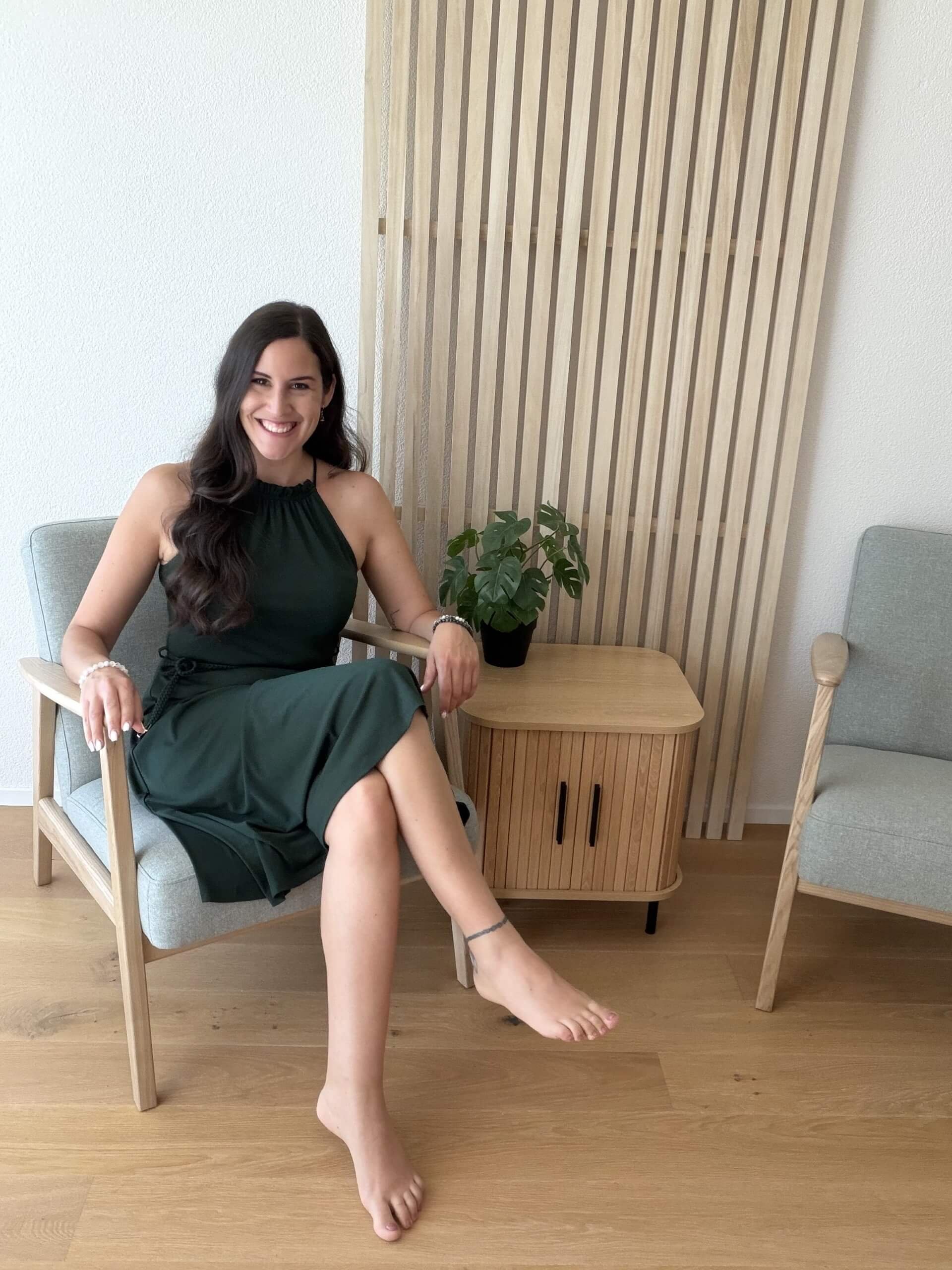 Eine junge Frau mit dunklen, lockigen Haaren sitzt lächelnd in einem modernen, Holz und Stoffstuhl, der auf einem Holzfußboden steht. Sie trägt ein dunkles, ärmelloses Kleid und ist entspannt, mit einer Hand auf dem Armlehnen und einem Bein über das andere geschwungen. Im Hintergrund befindet sich ein dekoratives Holzwandpaneel und eine Topfpflanze auf einem kleinen Holztisch.