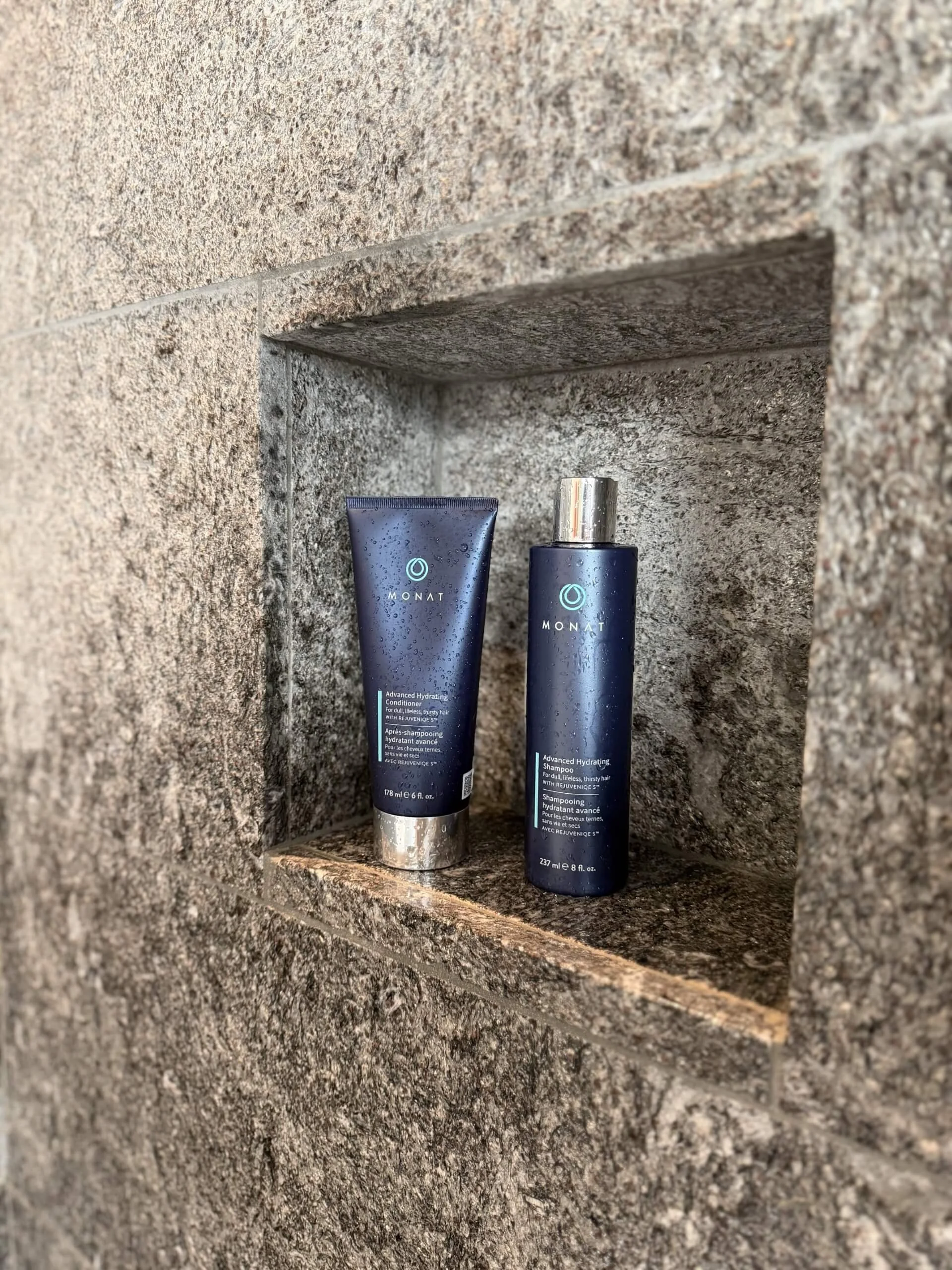 Zwei Monat-Haarpflegeprodukte, eines Shampoo und eines Conditioner, stehen in einem Wandregal aus Stein.