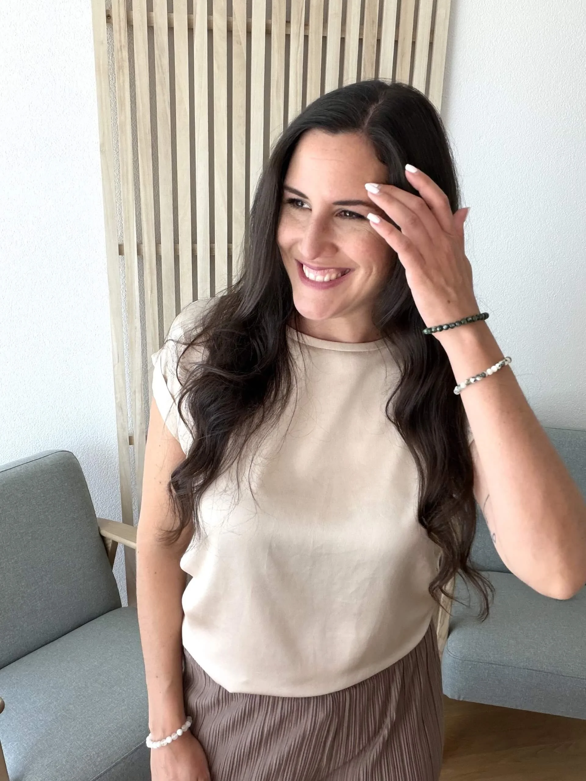 Eine lachende Frau mit langen braunen Haaren, die an der Seite hochgesteckt sind, sitzt in einem Wohnzimmer. Sie trägt einen hellen Oberteil, eine Armband aus Perlen am rechten Handgelenk und einen weiteren Armband aus schwarzen und weißen Perlen am linken Handgelenk. Sie hat eine Hand an der Stirn und blickt nach rechts.
