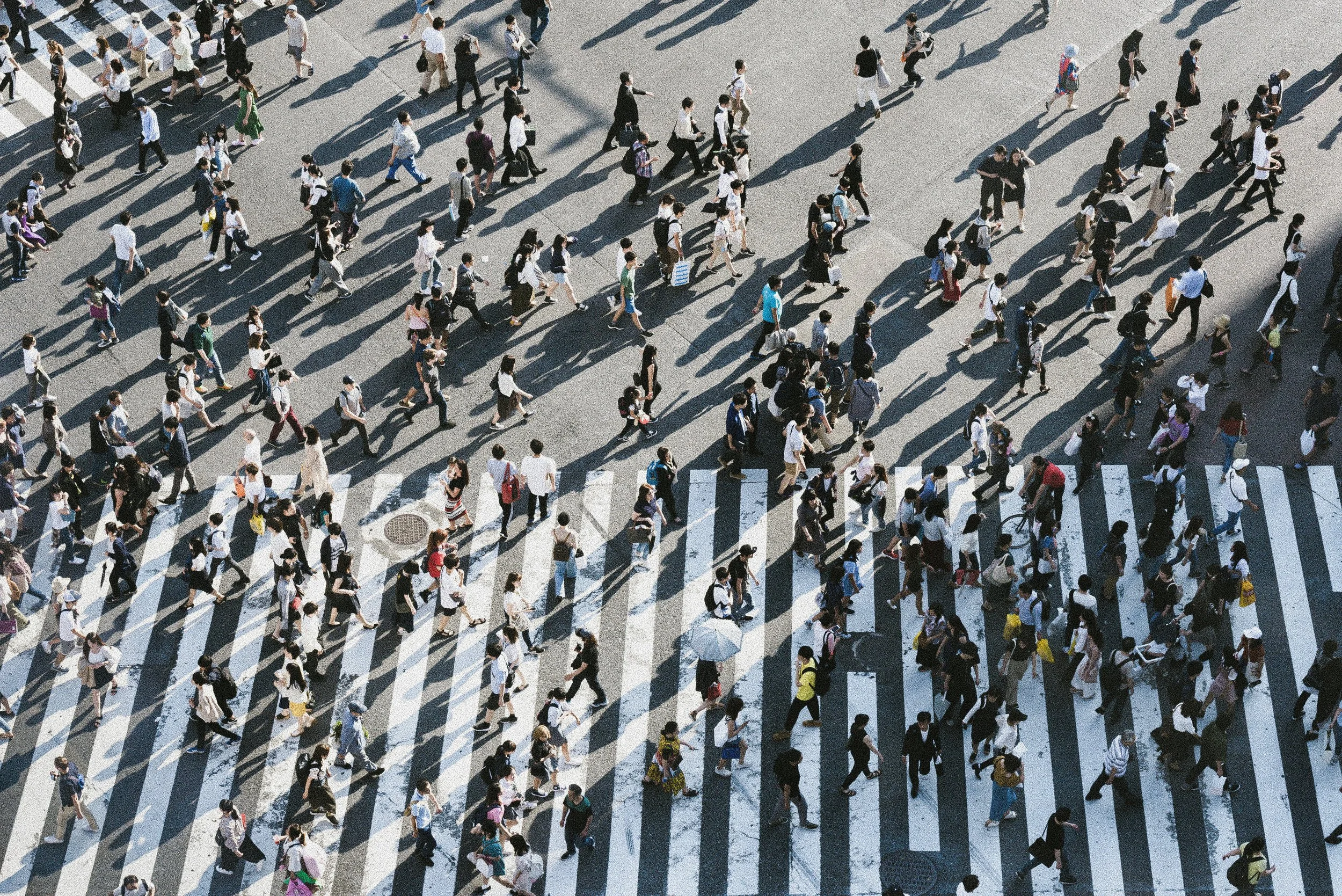 Aerial view of a busy crosswalk in an urban area with people walking in different directions, casting shadows on the pavement.