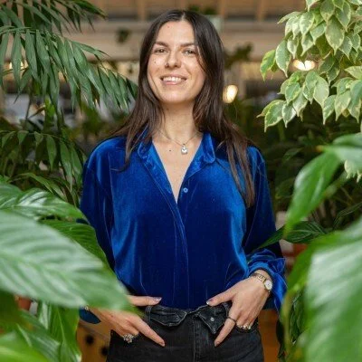 A woman with long dark hair smiling, wearing a blue blouse, standing among green indoor plants.