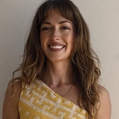 A woman with wavy brown hair smiling, wearing a yellow patterned top, standing in front of a plain wall.