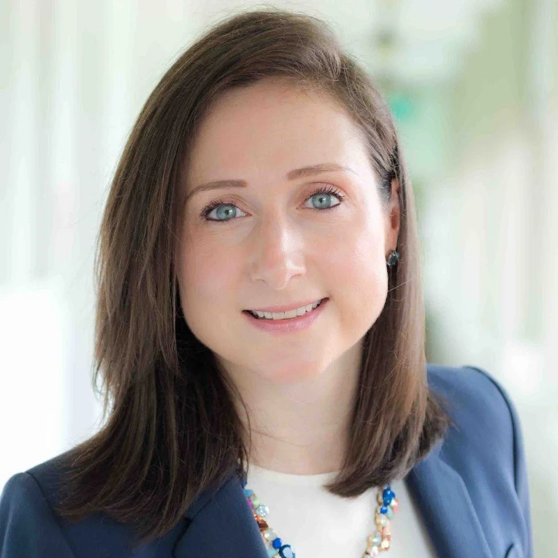 Portrait of a smiling woman with blue eyes and brown hair wearing a navy blazer and a colorful beaded necklace in a bright, outdoor setting.
