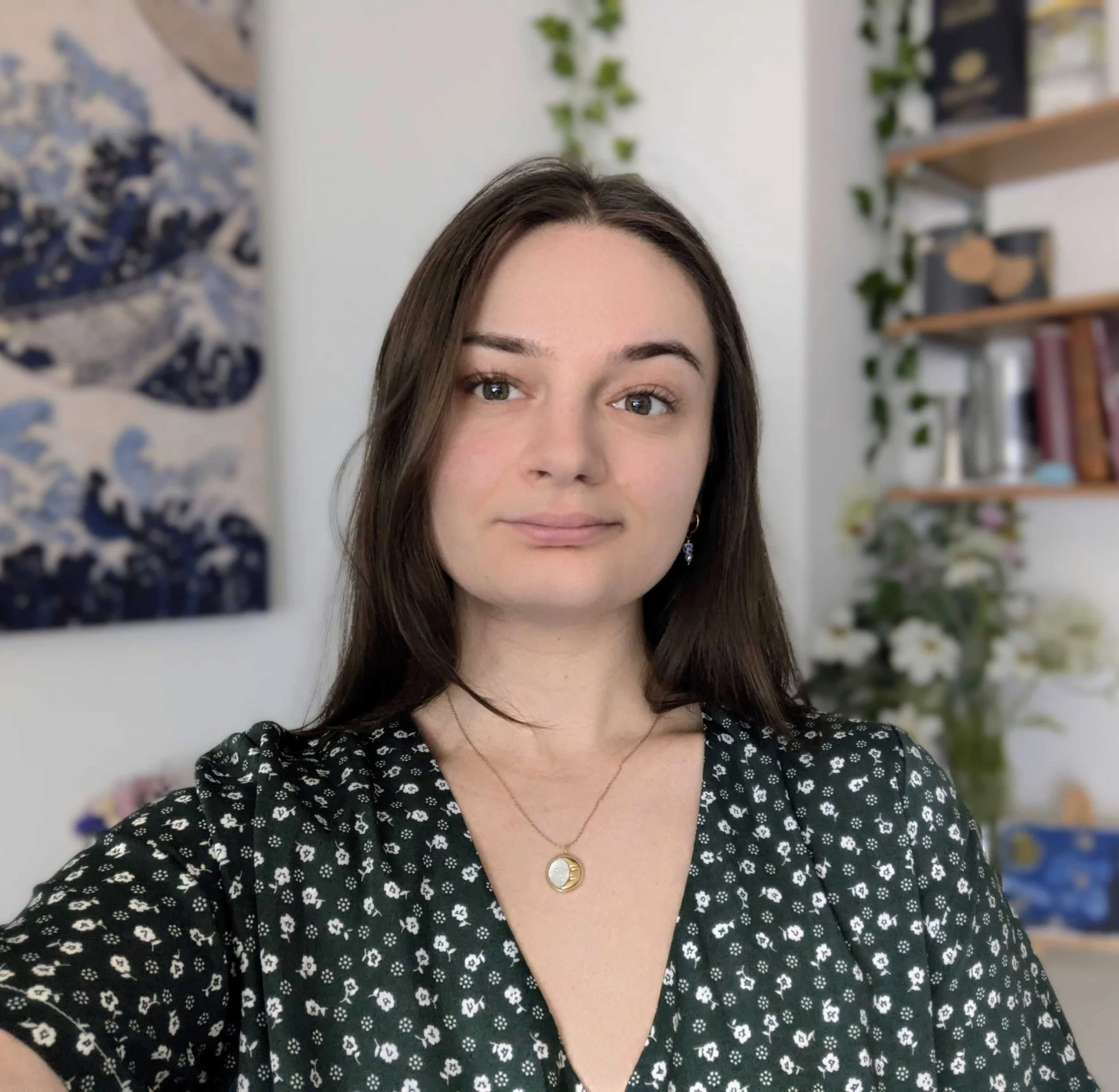 A woman with long brown hair wearing a black dress with white floral pattern, a gold necklace with a circular pendant, and earrings, standing in a room with a bookshelf and greenery in the background.