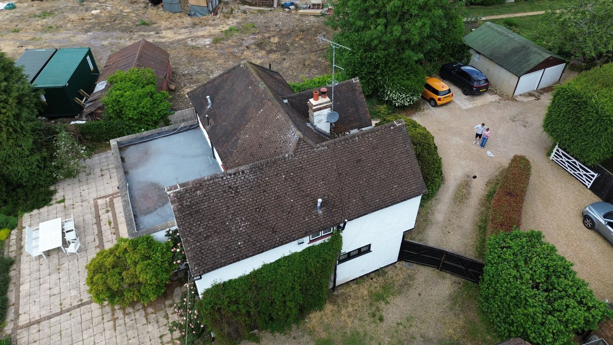 An aerial view of a backyard with multiple houses, trees, and parked cars. Two people are standing together near the center of the yard.