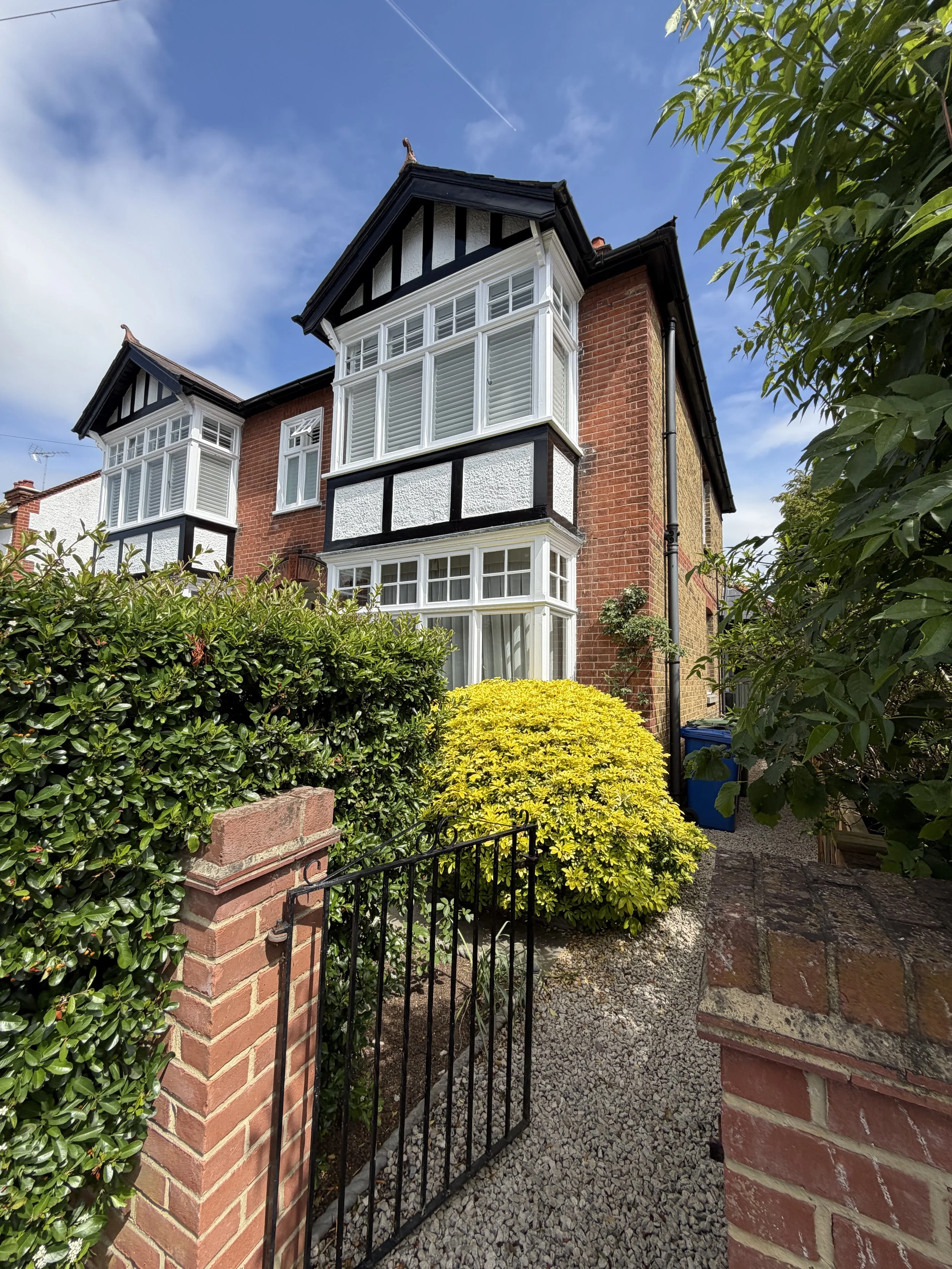 Front view of a brick house with large bay windows, a small gated entrance, and surrounded by green bushes and trees under a blue sky.