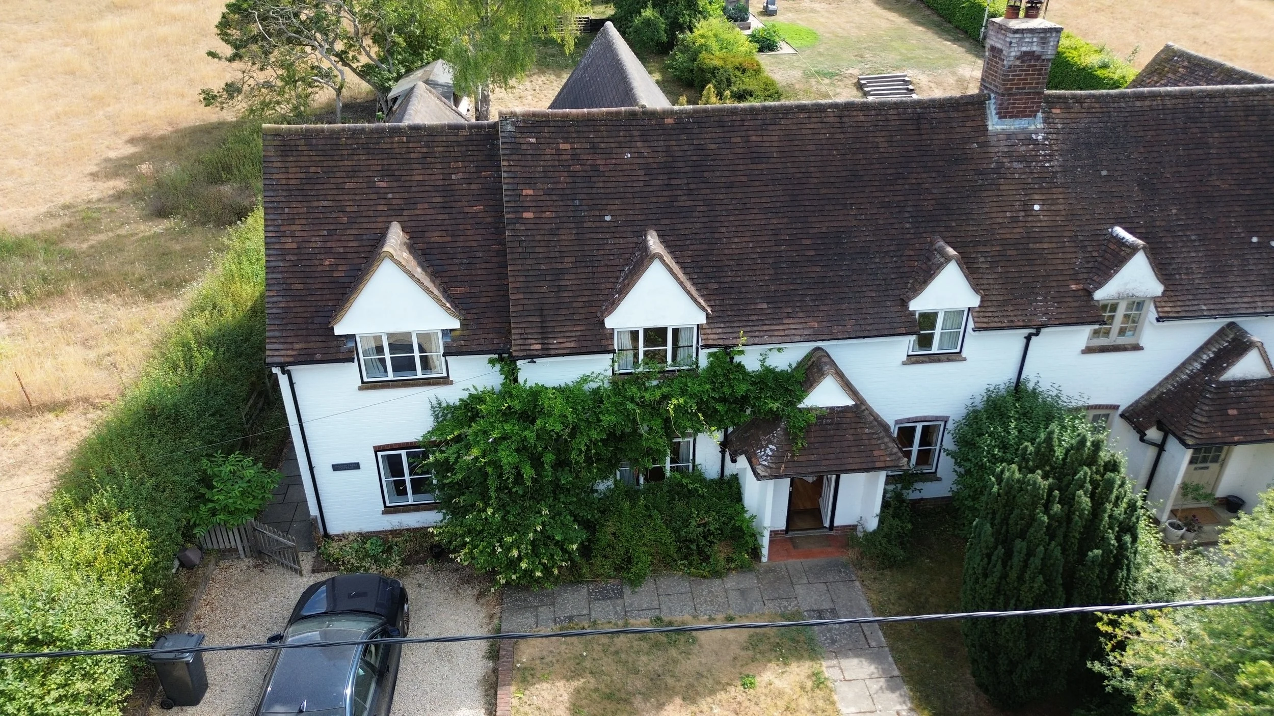 An aerial view of a white house with a brown tiled roof, surrounded by trees, green shrubs, and a small front yard, with a black car parked in a gravel driveway.