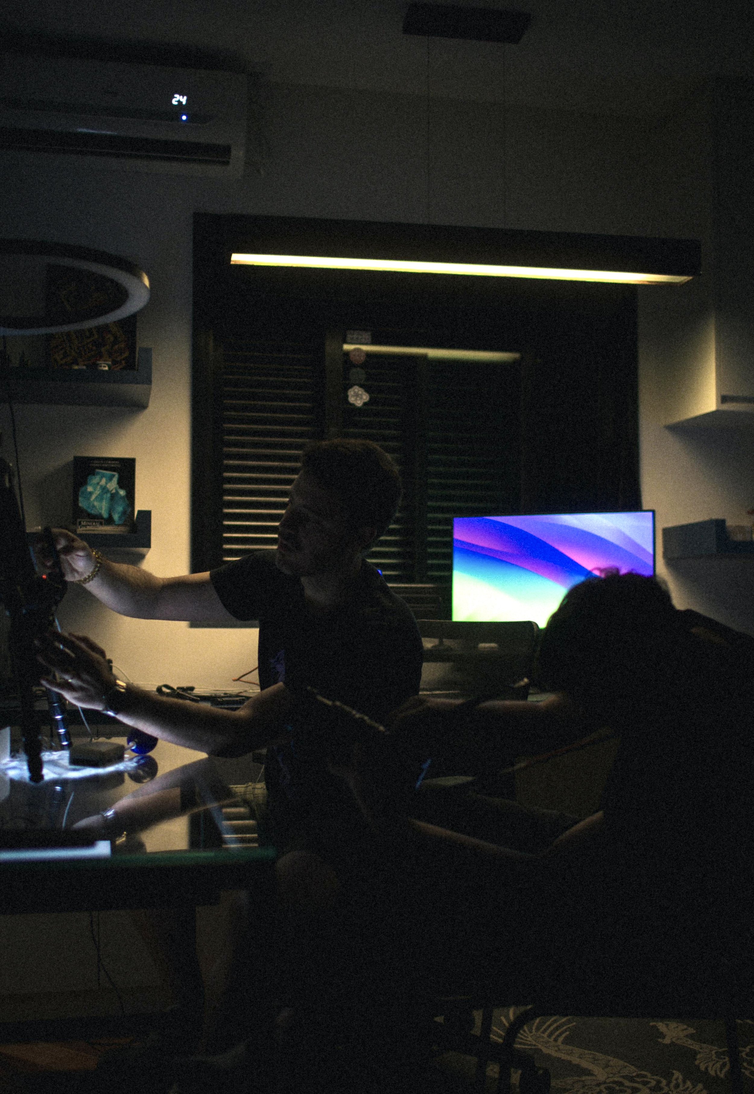A man in a dark room appears to be working with audio equipment. There is a television or computer monitor displaying a colorful screen, and some shelves with decorative items. A split air conditioning unit is mounted on the wall.
