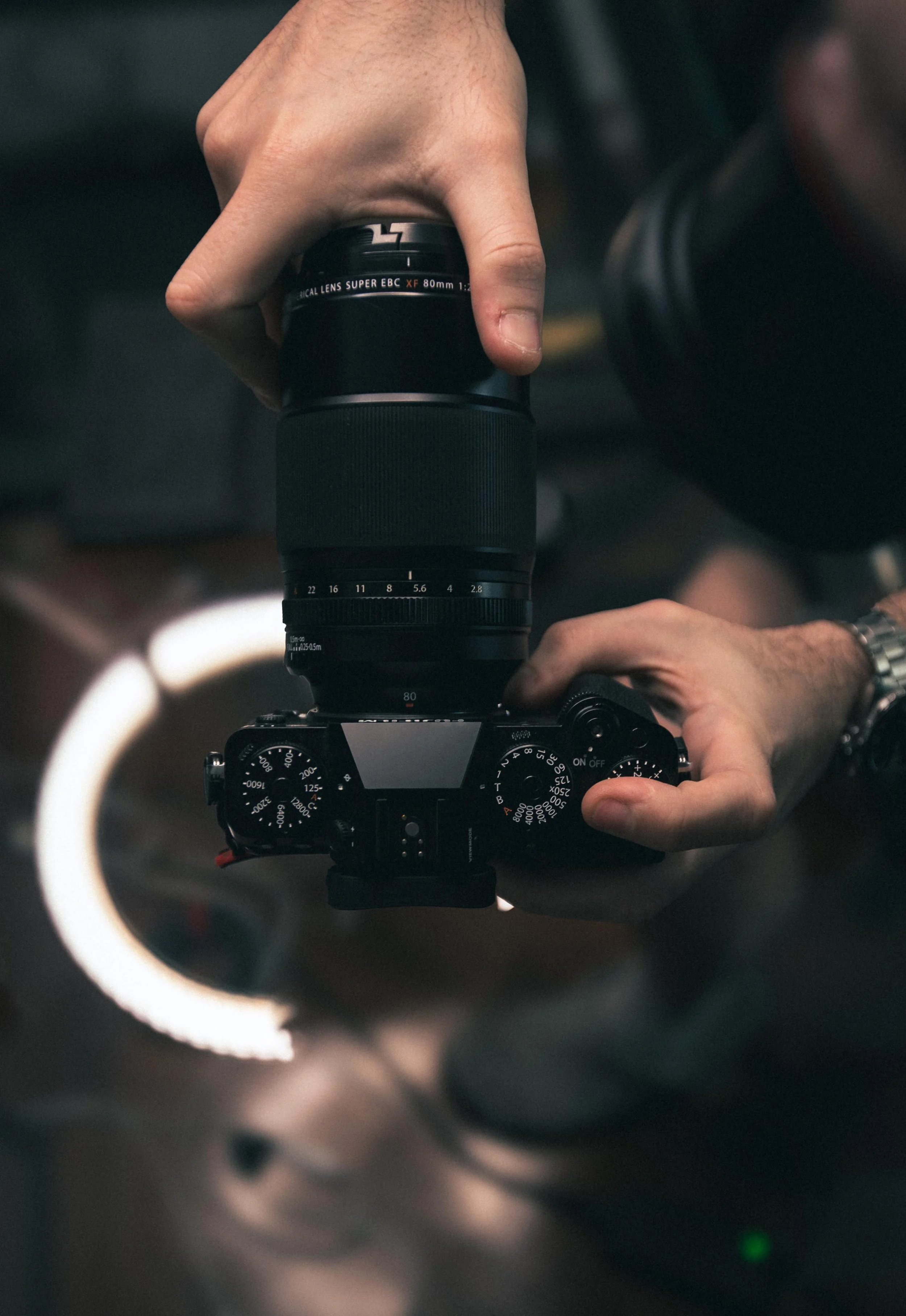 Person holding a camera with a lens, seen through a ring light, with their finger on the camera's shutter button.
