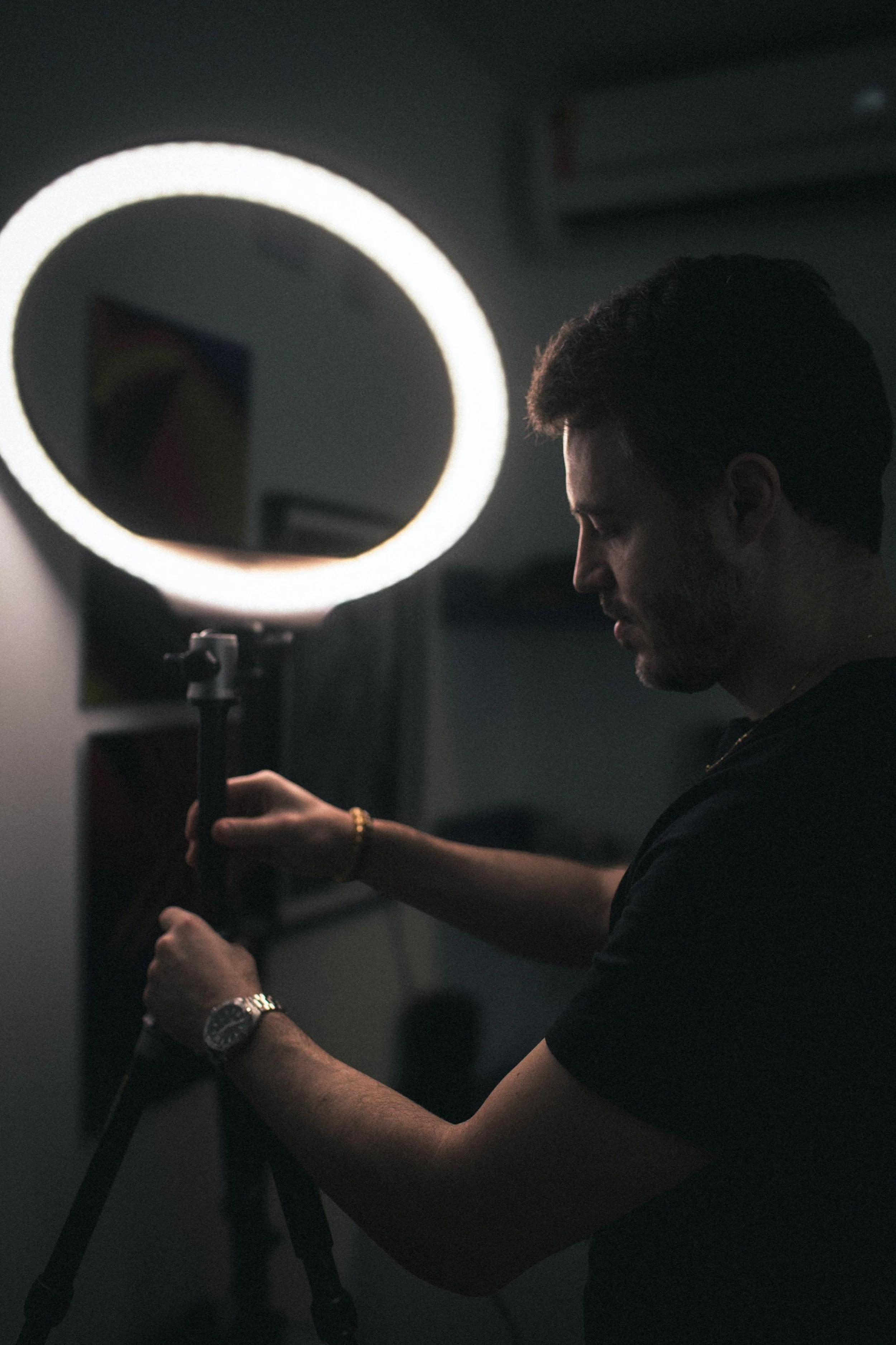 A man with dark hair and a beard, wearing a black shirt and a wristwatch, is adjusting or fixing something on a piece of equipment in a dimly lit room. The equipment has a circular light source.