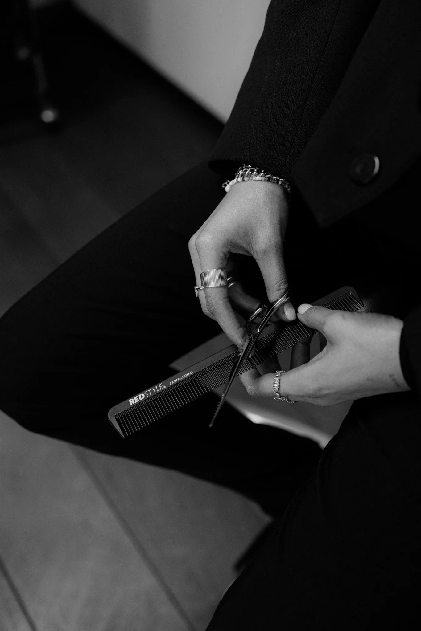 Schwarz-Weiß-Fotografie einer Person, die frisiert, hält einen Kamm und eine Haarschneideschere in den Händen, trägt Ringe und Armbänder, trägt einen schwarzen Blazer, sitzt auf einem Stuhl, im Hintergrund ein dunkler Raum.