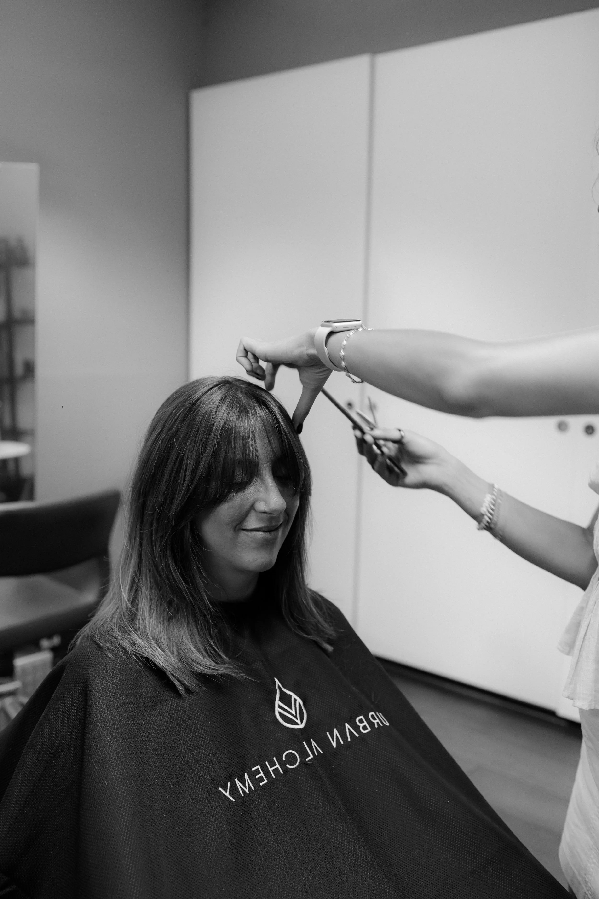 Eine Frau mit braunen Haaren sitzt in einem Friseurstuhl, während eine Friseurin ihre Haare schneidet. Die Szene findet in einem Friseursalon statt.