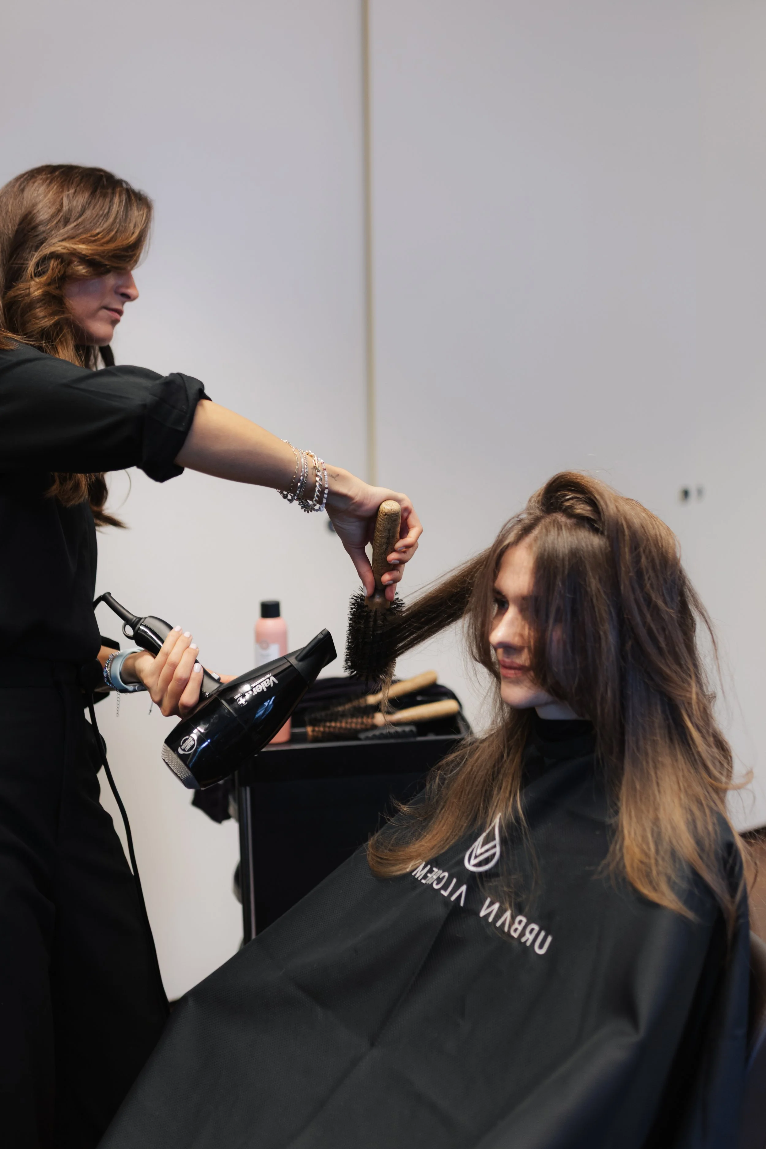 Friseur beim Haarstyling einer Frau in einem Friseursalon, mit Haartrockner in der Hand, Frau sitzt auf einem Stuhl mit Umhang.