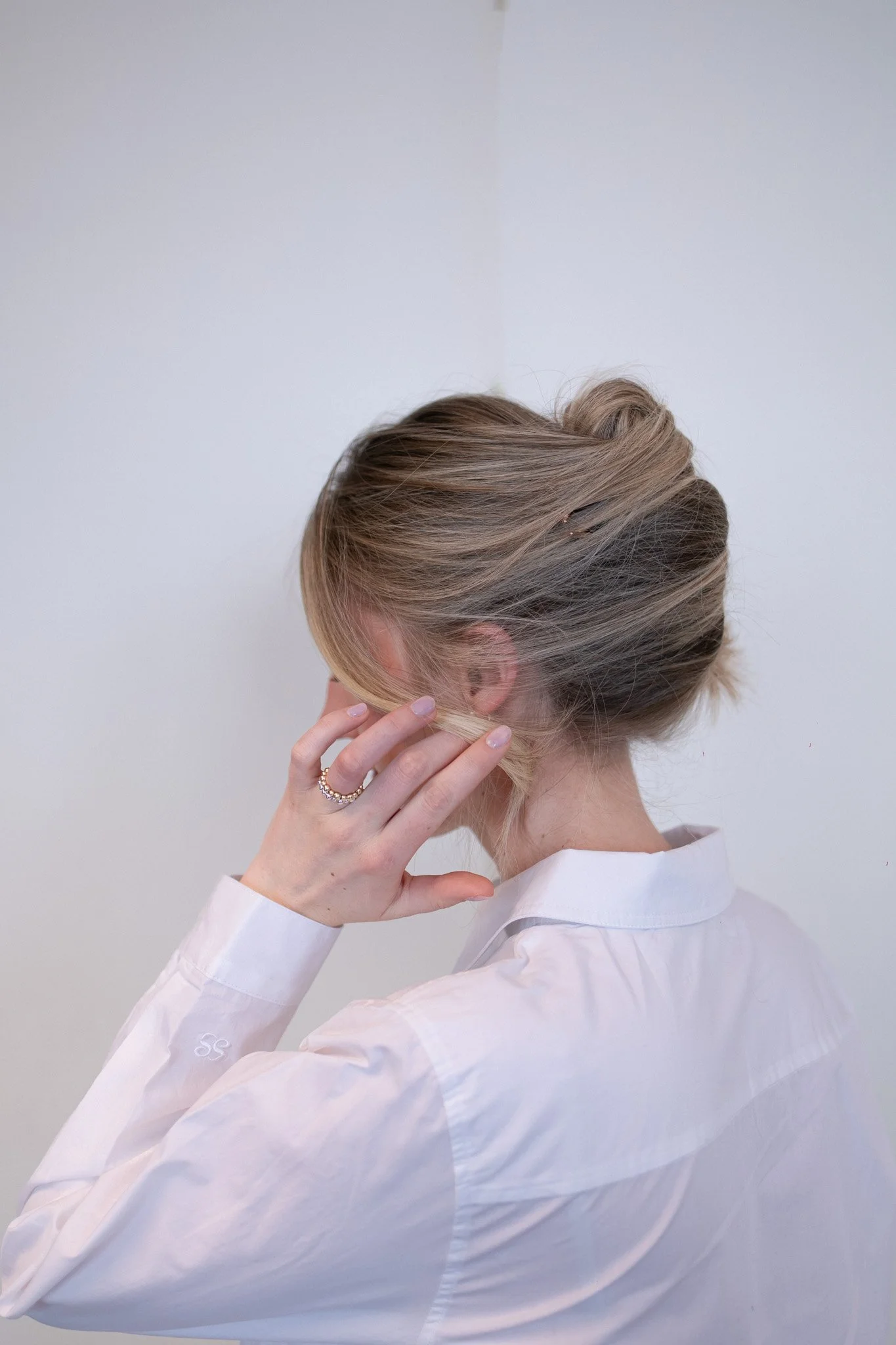 Eine Person mit blonden Haaren trägt ein weißes Hemd und hält die Hand an das Ohr, während sie seitlich fotografiert wird, vor einem neutralen Hintergrund.