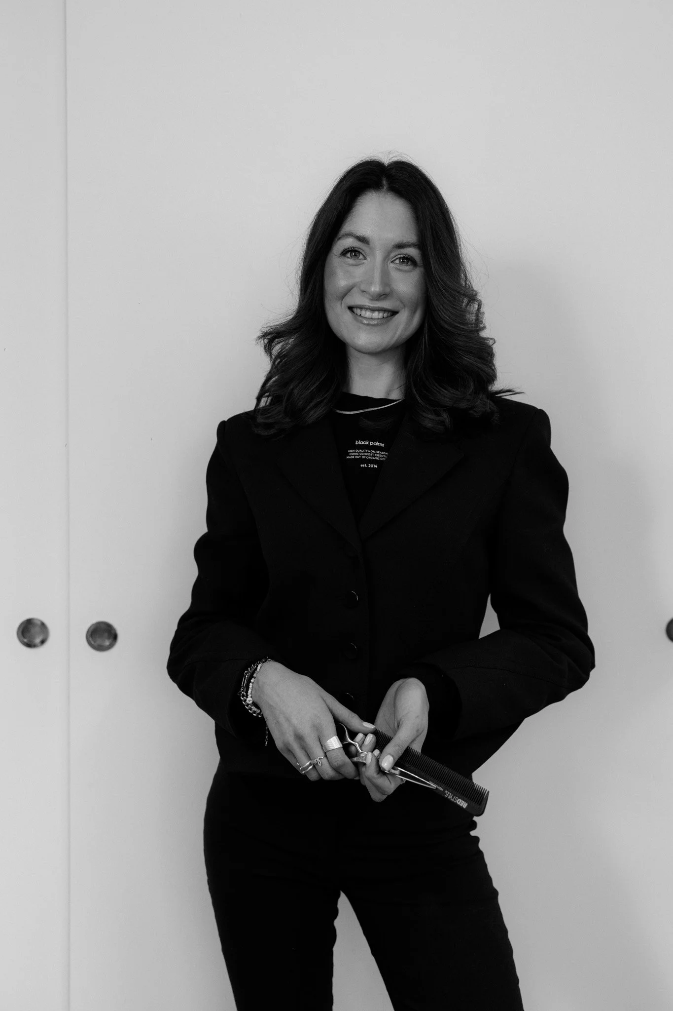 Schwarz-weiß-Fotografie einer lächelnden Frau mit dunklen, gewellten Haaren, die eine schwarze Jacke trägt, vor einer weißen Wand. Sie hält ein Maßband in den Händen.