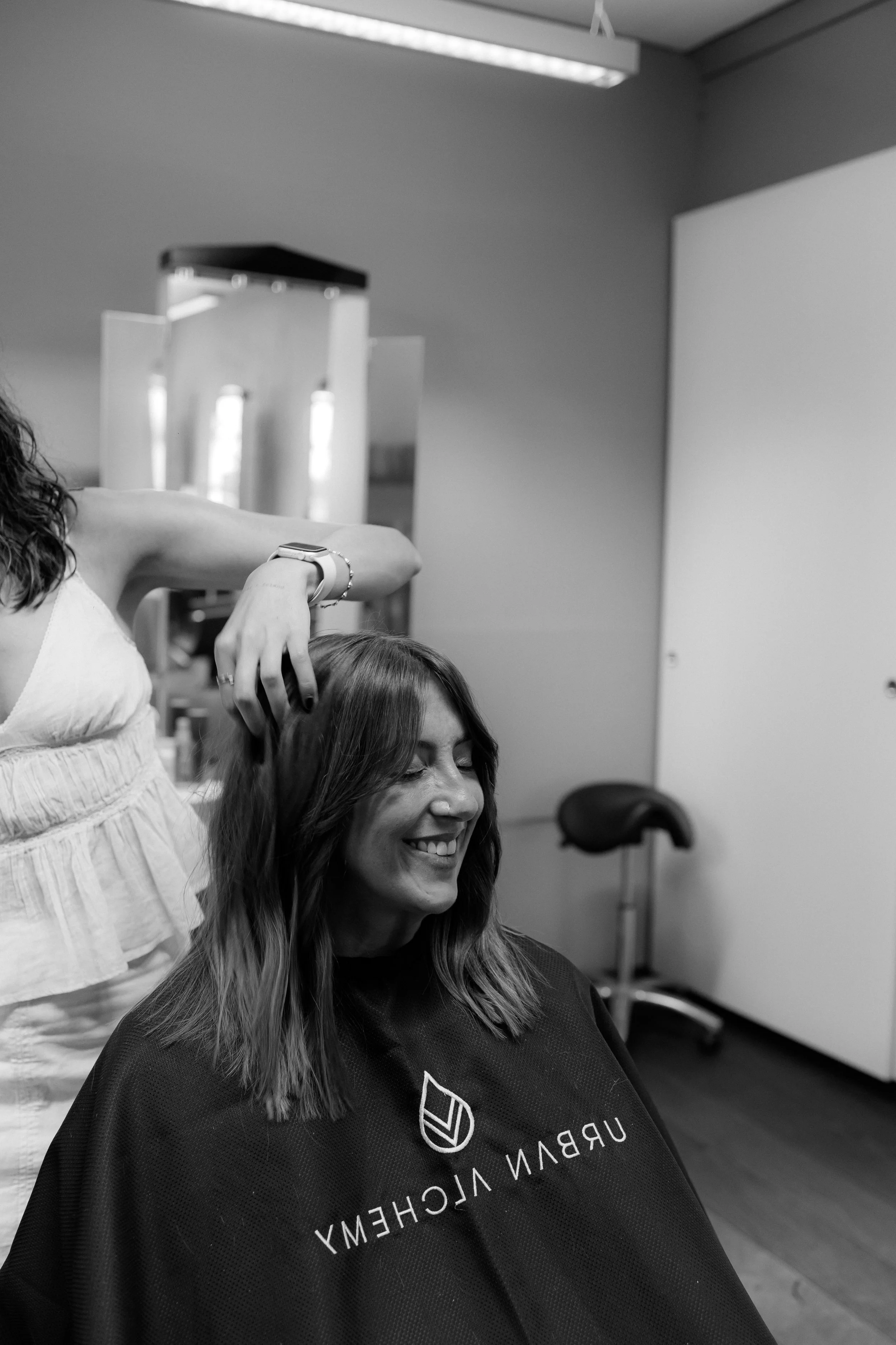 Eine Frau mit braunen Haaren, die lächelnd auf einem Frisiertisch sitzt, während eine andere Frau mit dunklen Haaren sie frisiert.