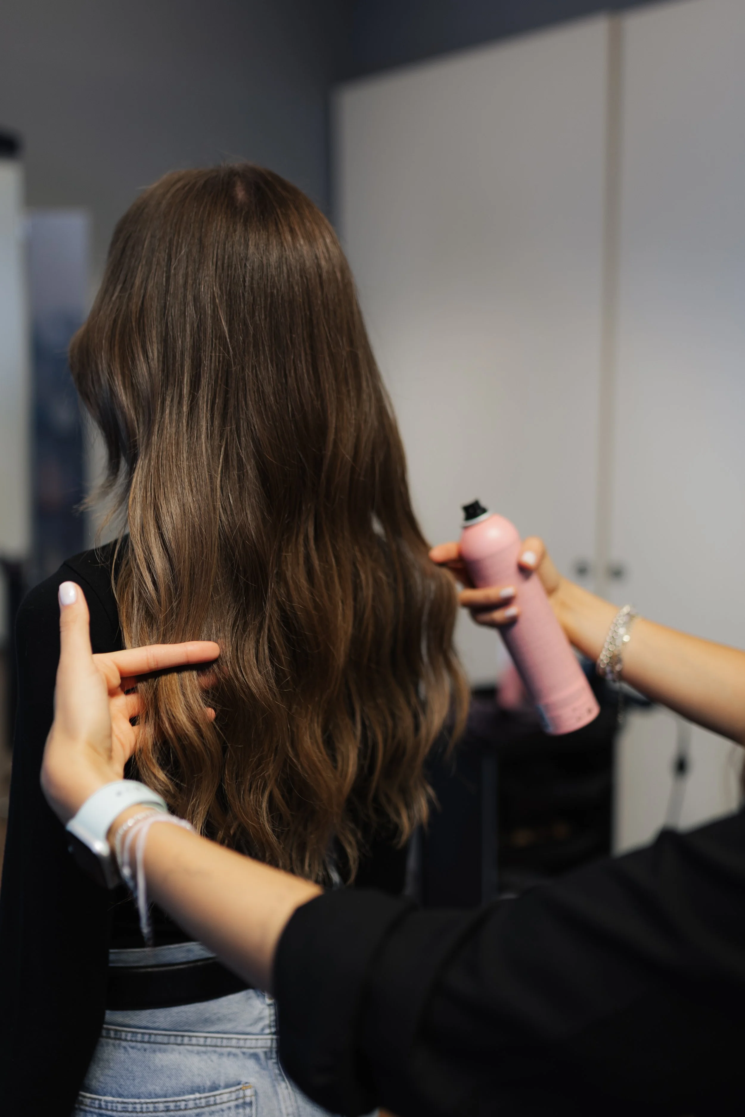 Frau mit braunen, gewellten Haaren wird in einem Salon gestylt, während eine Stylistin ein Haarstyling-Produkt auf ihren Haaren sprüht.