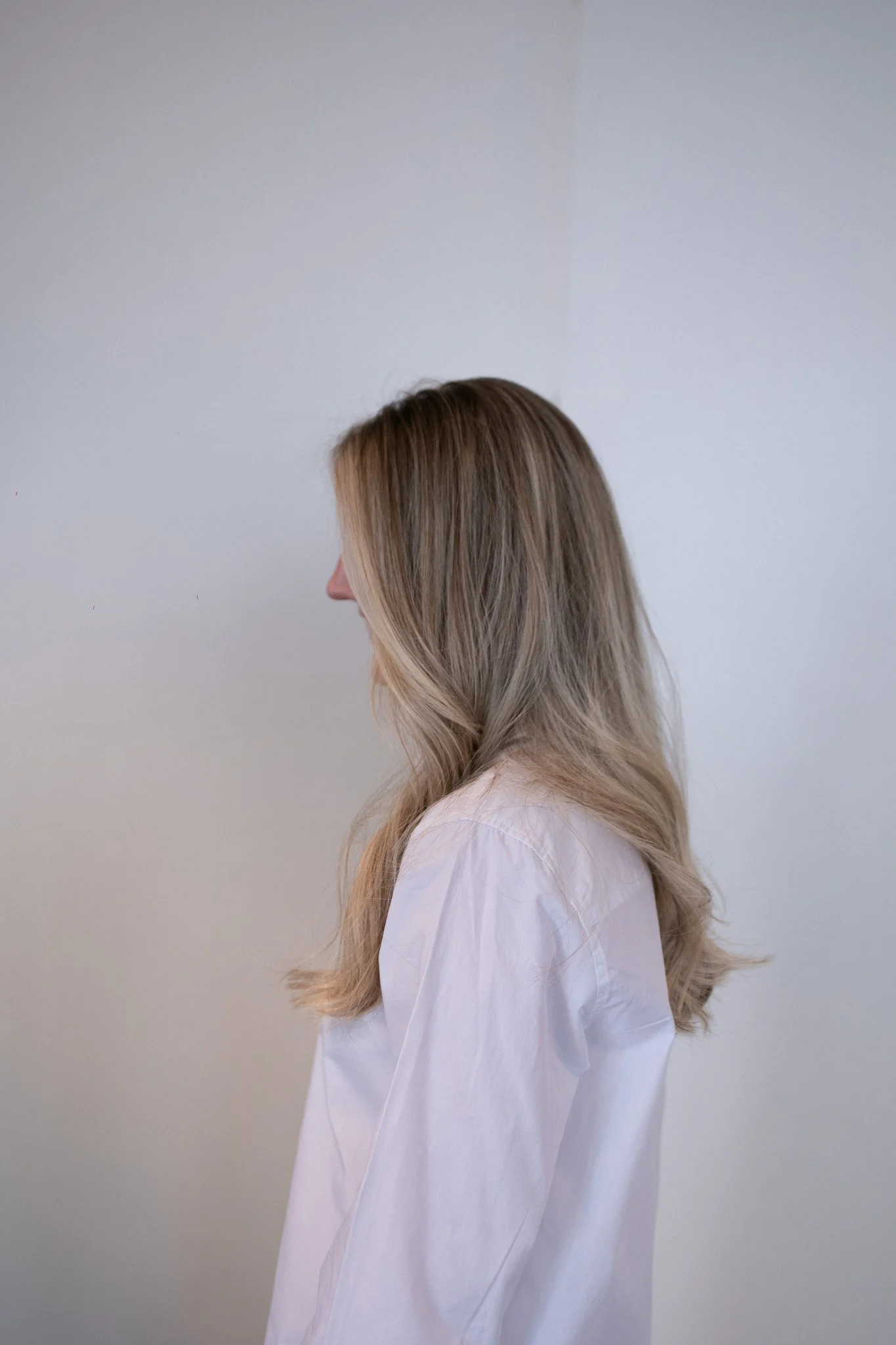 Seitenansicht eines Frauenköpfchens mit blonden, welligen Haaren und weißem Hemd gegen eine schlichte graue Wand.