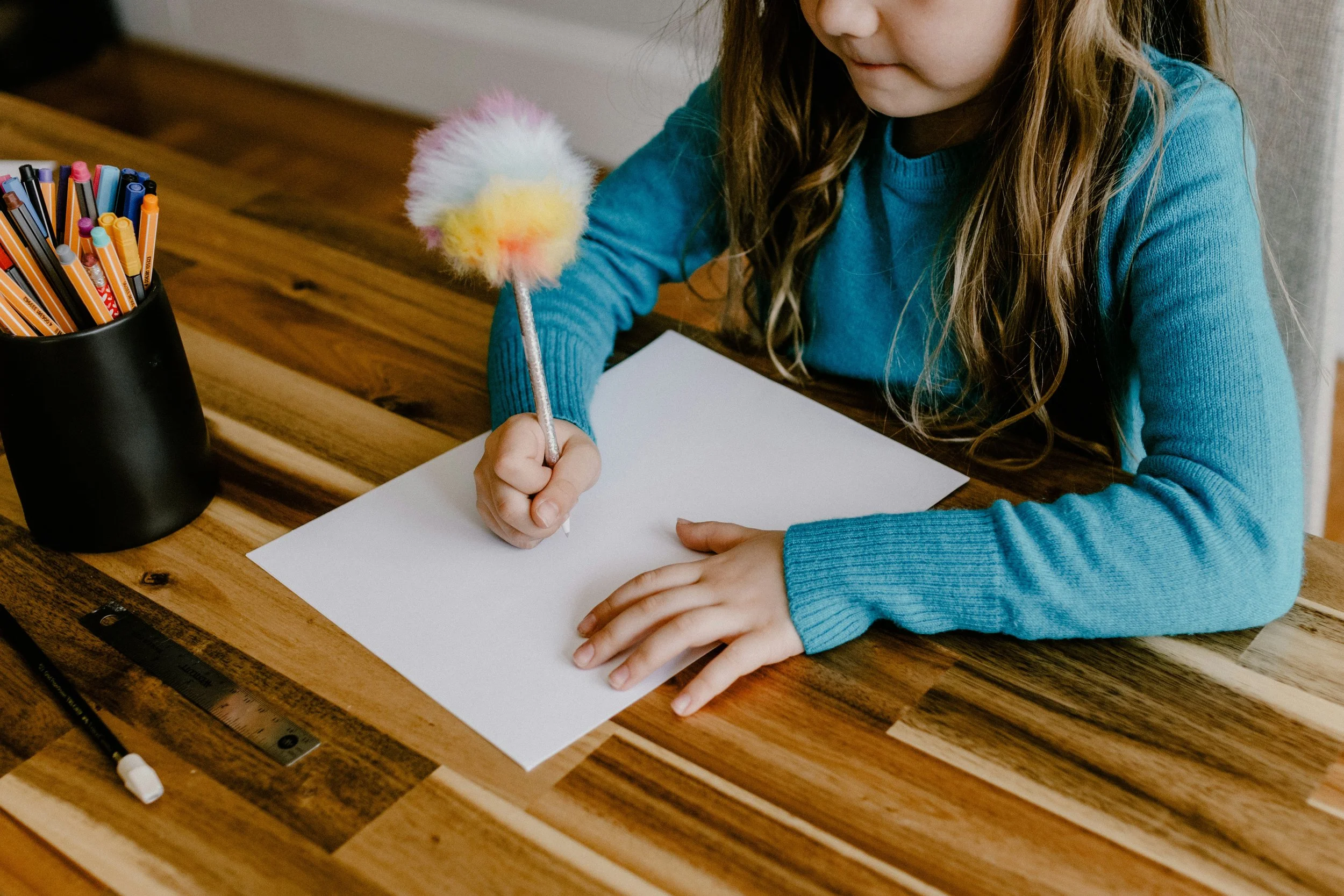 A young girl with long wavy brown hair wearing a blue sweater, sitting at a wooden table, holding a colorful fluffy cotton ball with a stick, preparing to write or draw on a blank white sheet of paper.