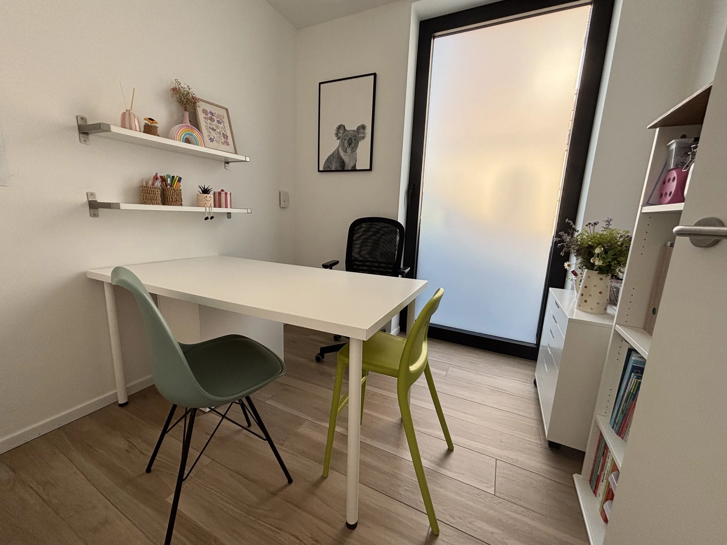 A corner of a room with a white table, three colorful chairs, two floating shelves with decorative items, a framed picture of a koala, a window with frosted glass, a bookshelf with books and photos, and a small potted plant.