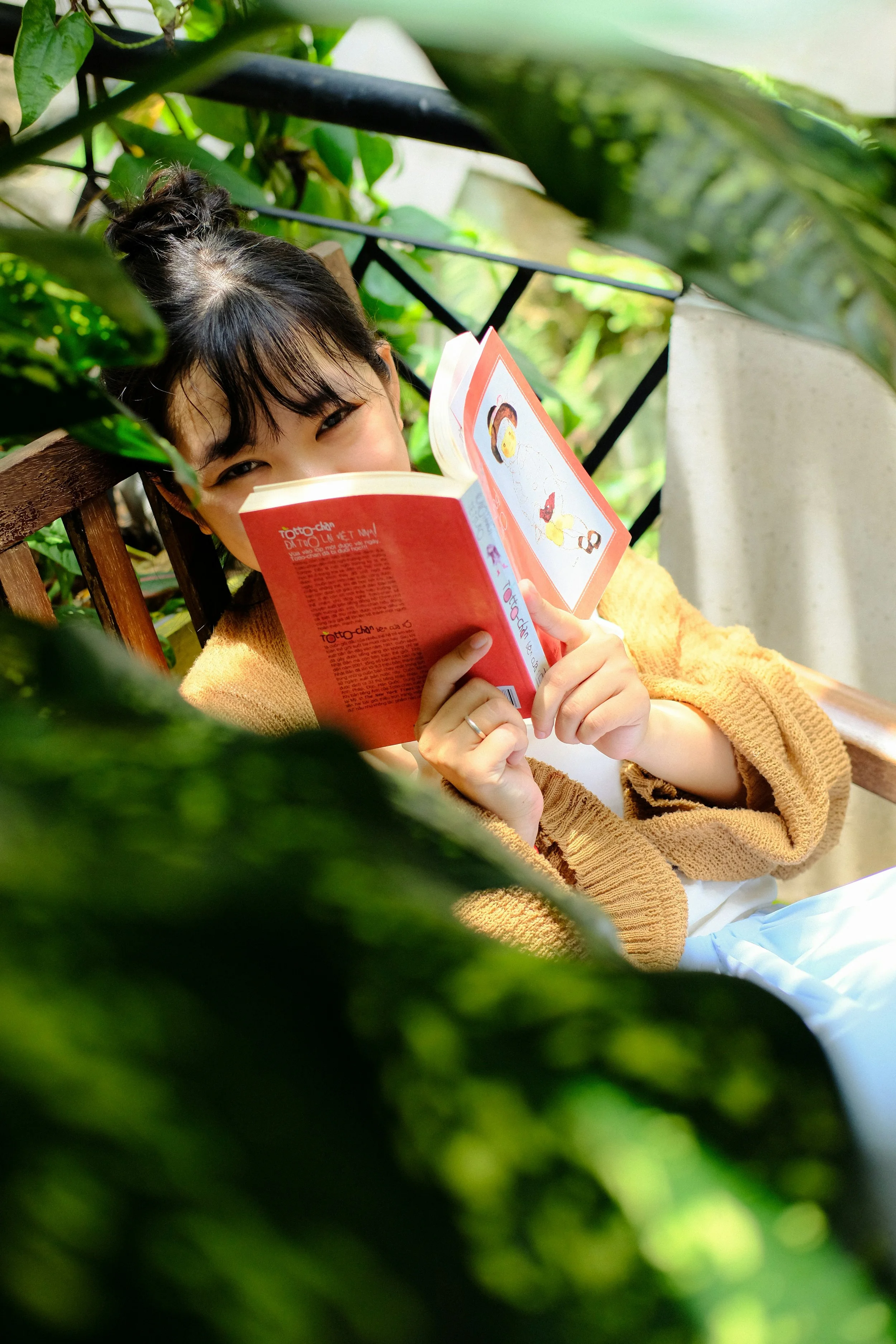 A young woman with dark hair in a bun, wearing a yellow sweater, sitting on a bench outdoors, partially hidden by green leaves, reading a red book.
