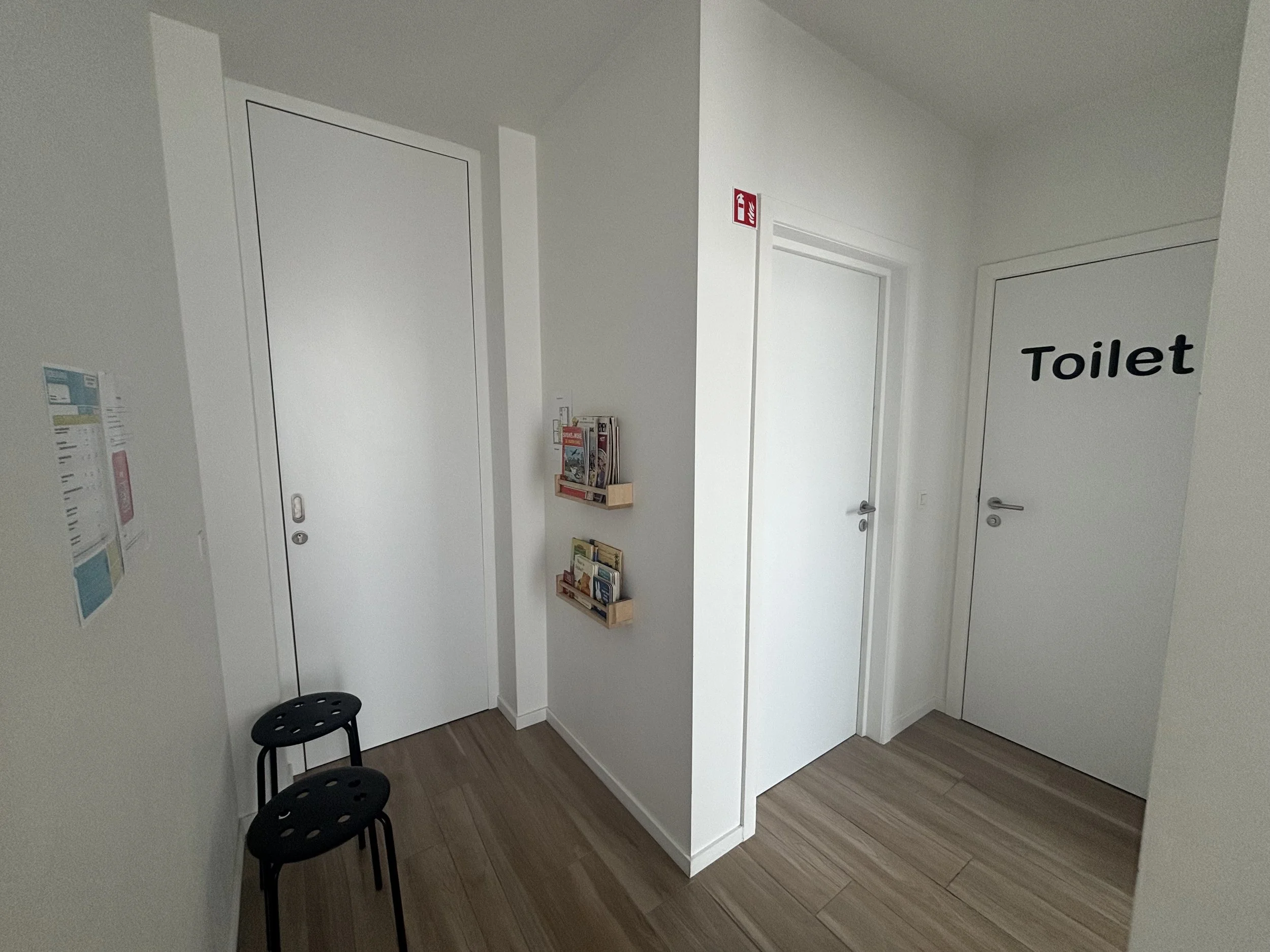 A corner of a room with white walls, two white doors labeled 'Toilet,' a small brochure rack attached to the wall, and three black chairs with round holes in the seat backs. The room has wooden flooring and a fire extinguisher sign on the wall.