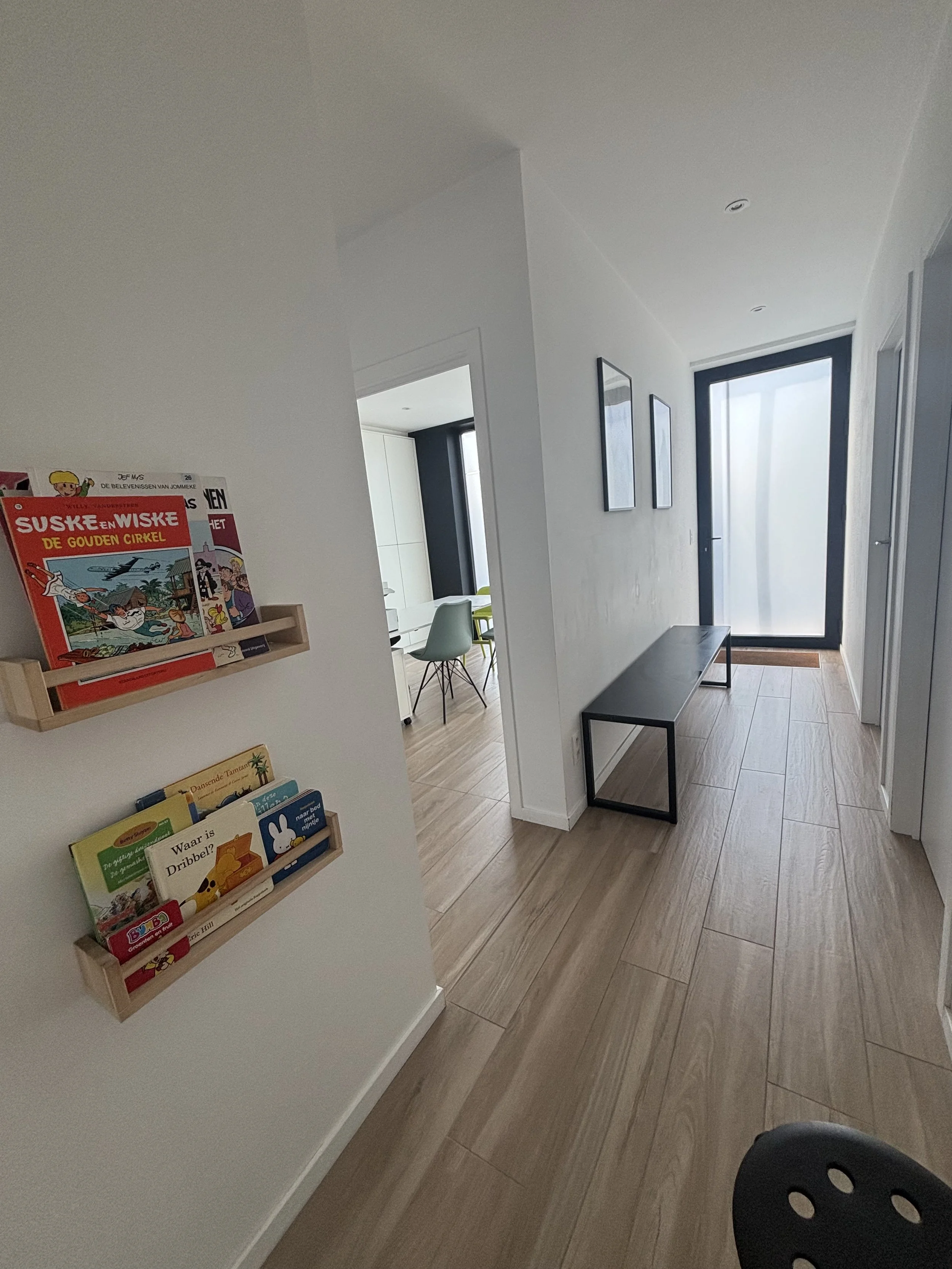 Interior view of a modern house hallway with light-colored wooden flooring, white walls, and black-framed glass doors. A small wooden bookshelf with children's books is mounted on the left wall. There are two framed pictures on the right wall, a blac