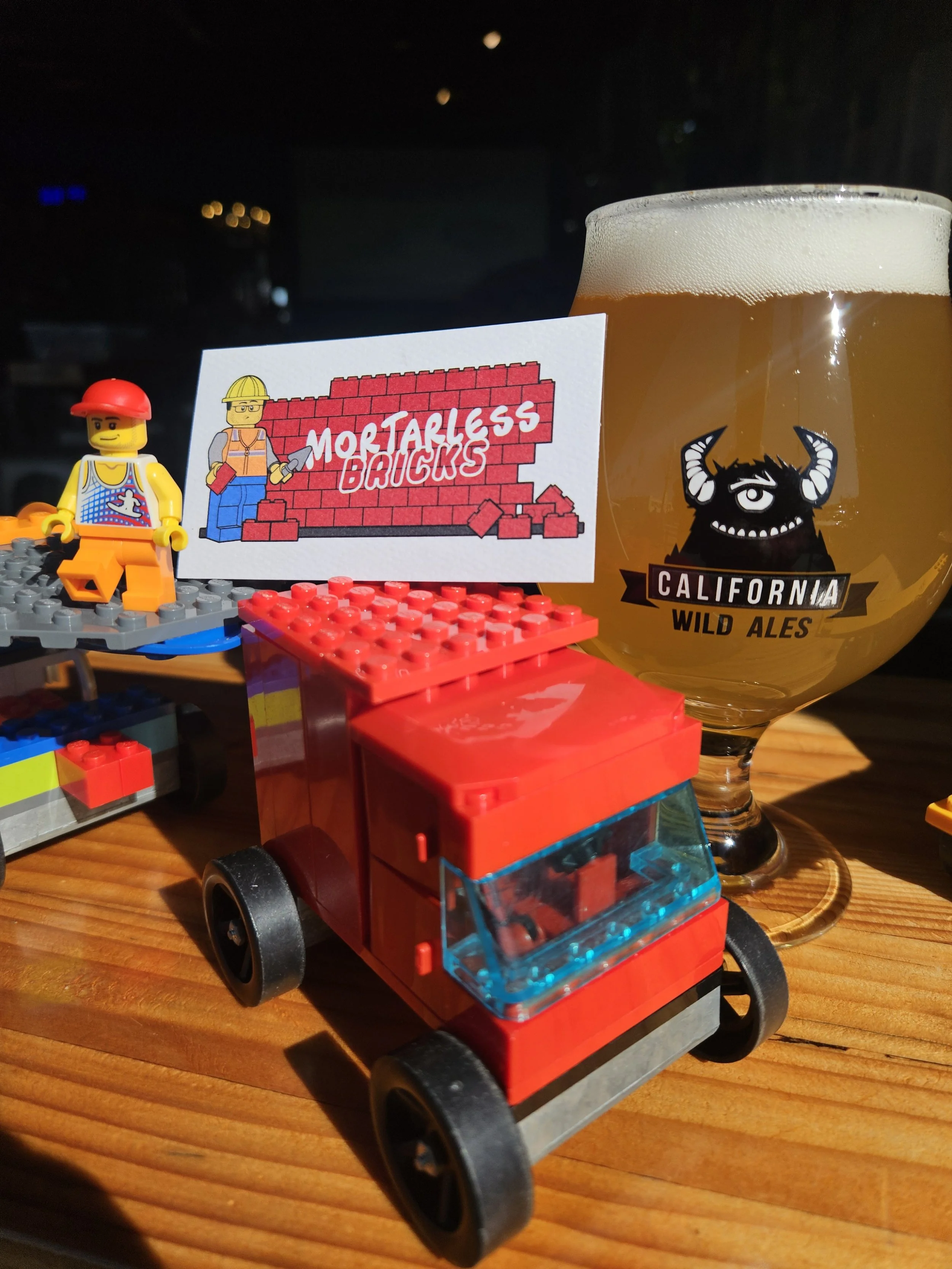 Lego construction worker figurine with a red hard hat, a card with a brick wall and the words 'Mortarless Bricks,' a glass of beer with 'California Wild Ales' logo, and toy bricks on a wooden surface.