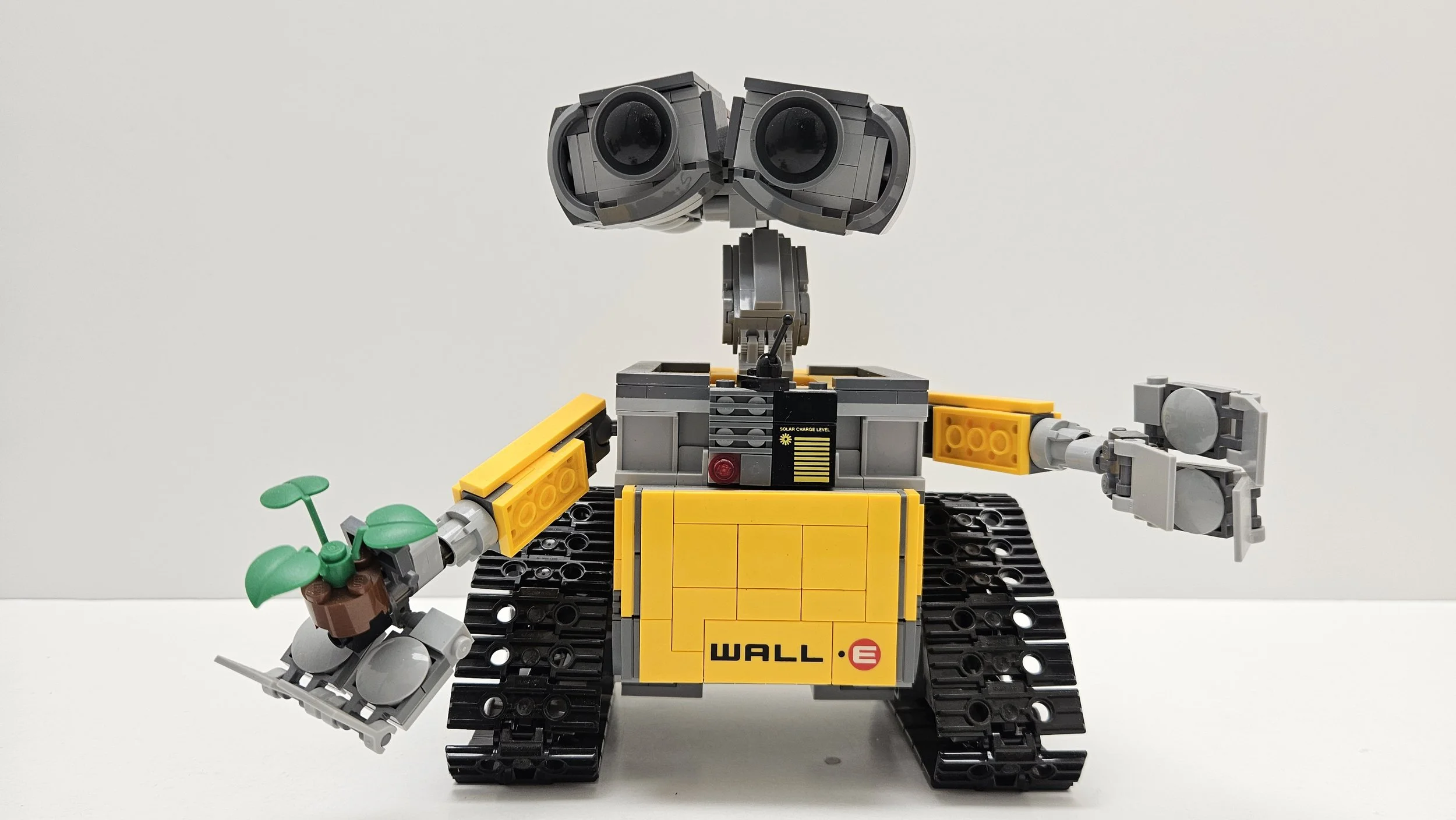A LEGO robot designed to resemble WALL-E from the animated film, holding a small plant in its left hand and standing on a white surface against a plain white background.