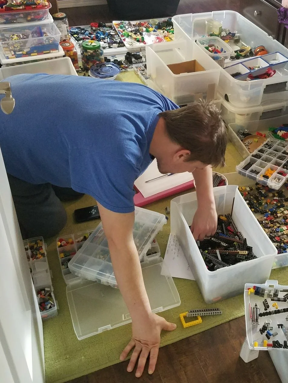 A person organizing LEGO bricks and pieces in plastic storage bins scattered on the floor, with additional containers full of LEGO toys and components in the background.