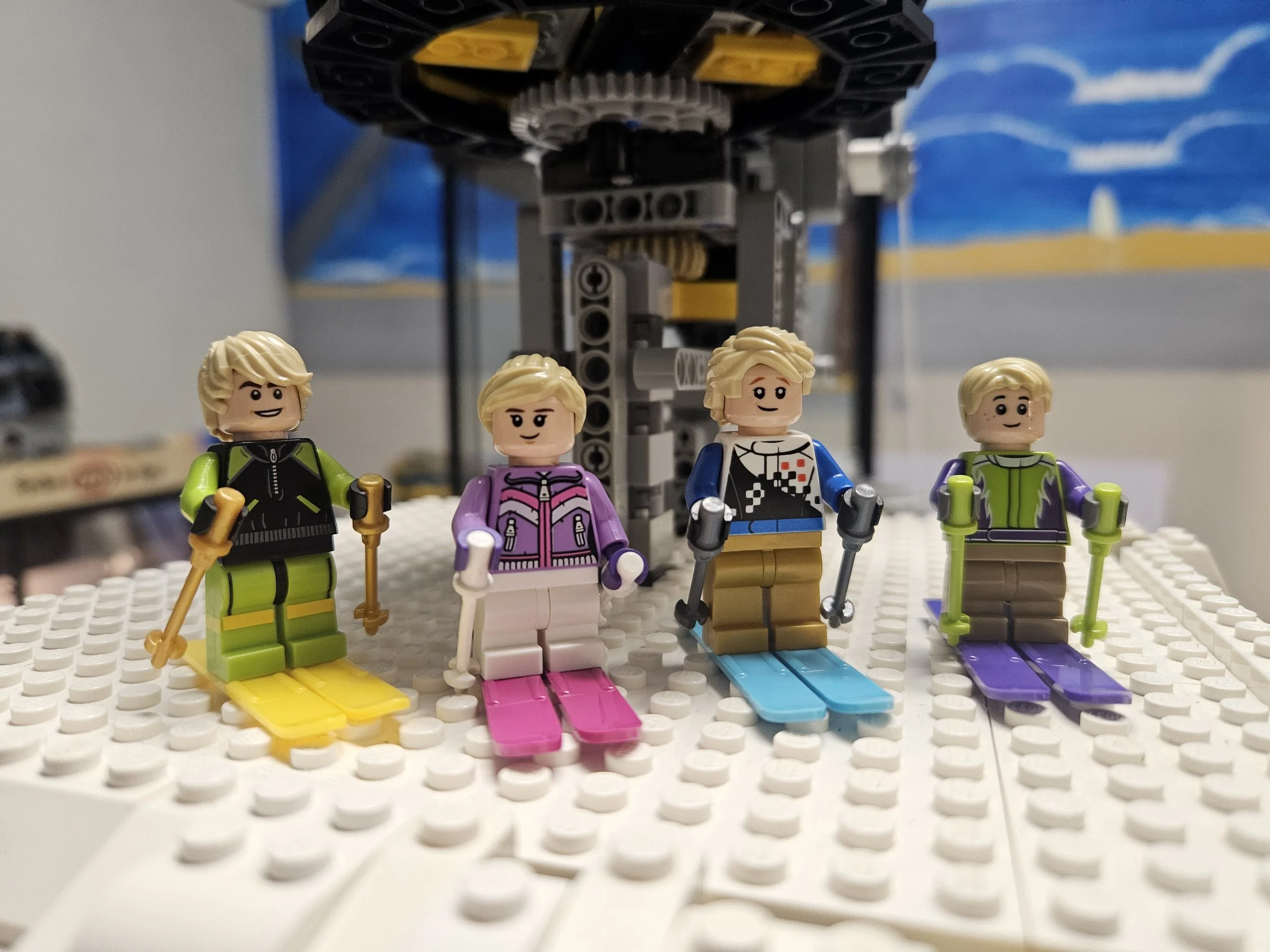 Four LEGO mini-figures dressed in colorful winter snowboarding outfits standing on a white LEGO baseplate with a black and yellow LEGO structure in the background and a painted wall with blue sky and white clouds.
