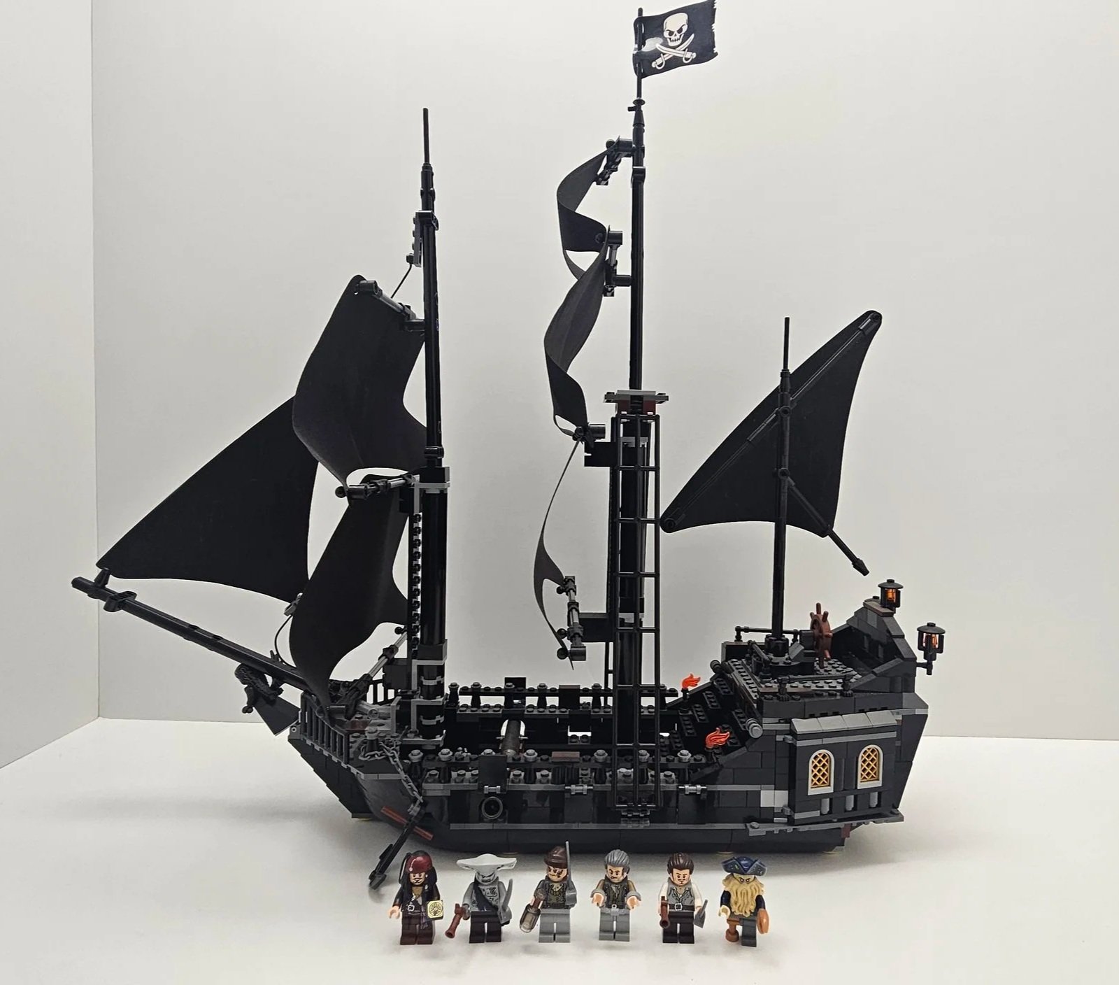 A LEGO pirate ship with black sails, a pirate flag with a skull and crossbones, and six LEGO minifigures in front.