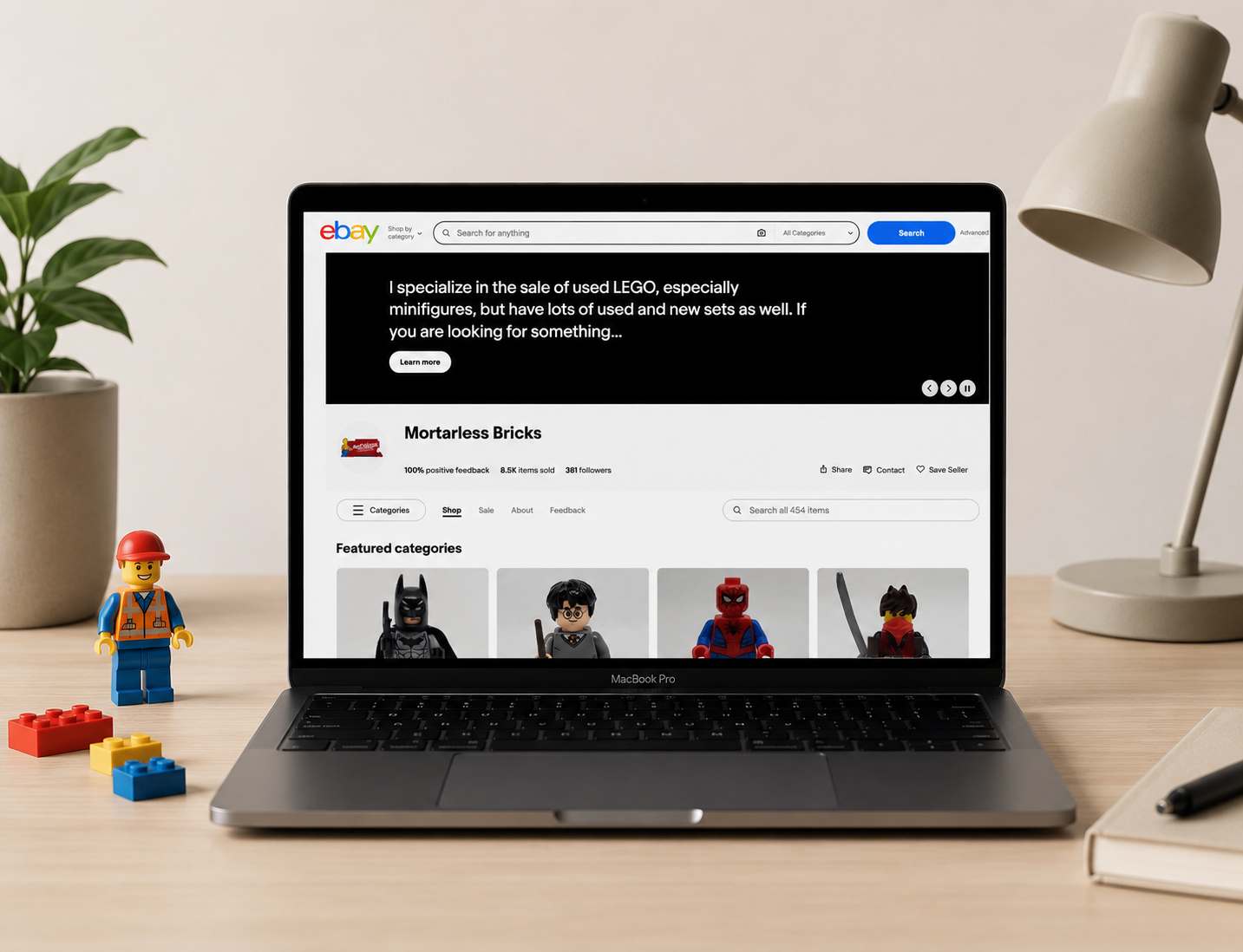 Laptop on desk displaying eBay page selling used LEGO minifigures and sets, with LEGO minifigures and LEGO bricks on desk, a small LEGO figure, a potted plant, and a desk lamp.