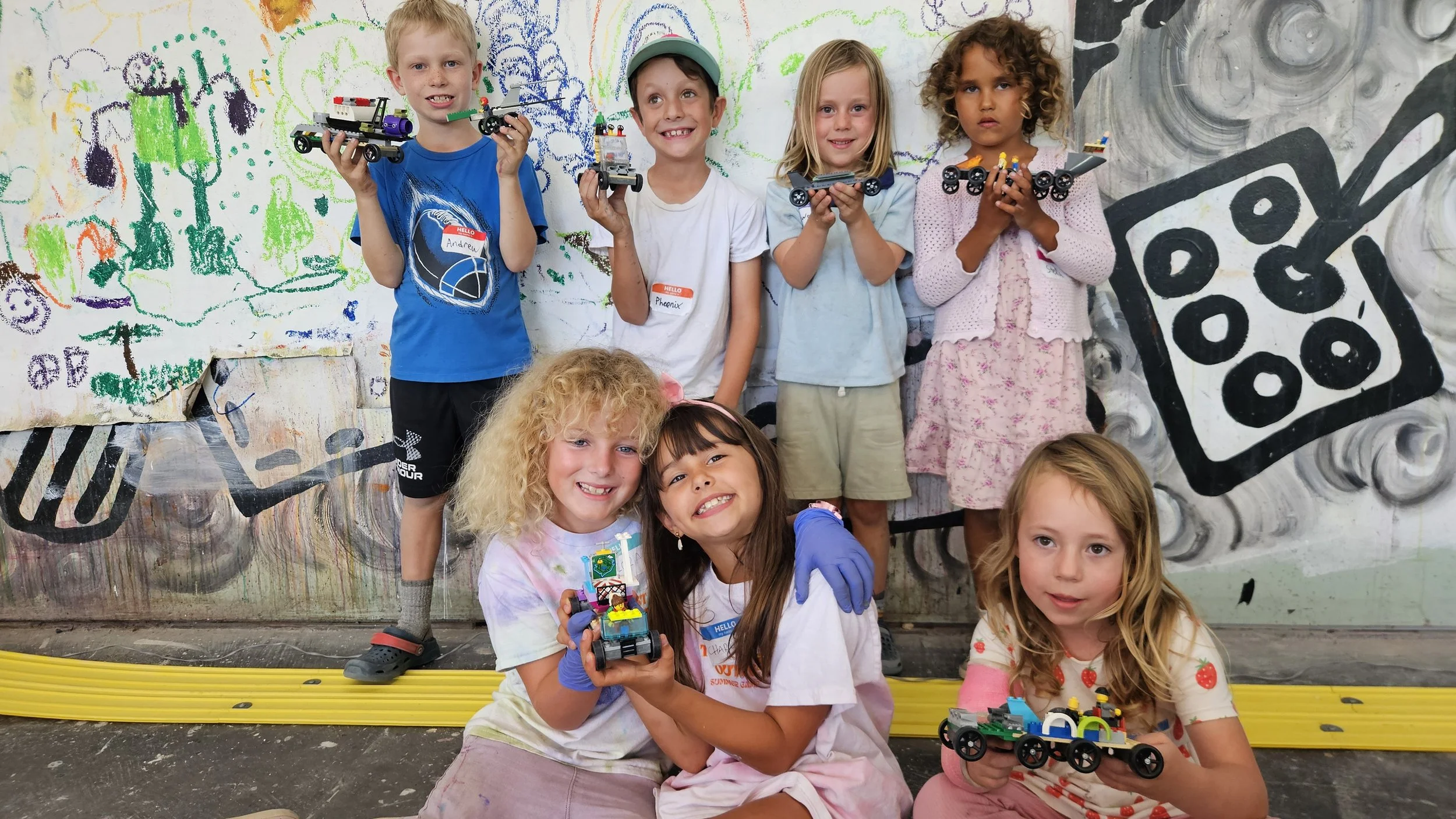 Group of children holding LEGO vehicles in front of a colorful graffiti wall with abstract designs and shapes.