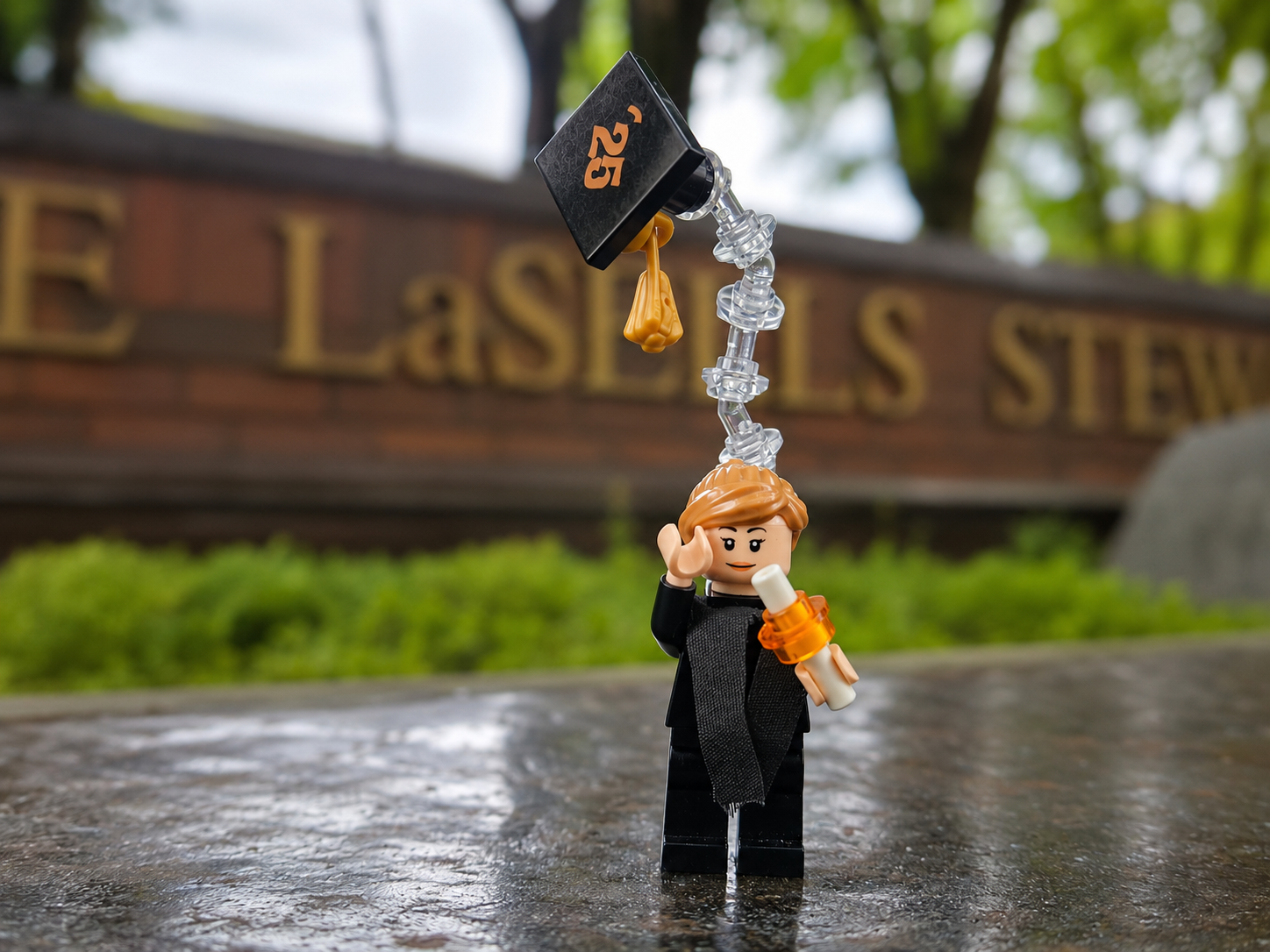 Lego figurine of a woman waving, holding a scroll, with a plastic arm extending above her head, set outdoors on a wet surface with greenery and a sign in the background.