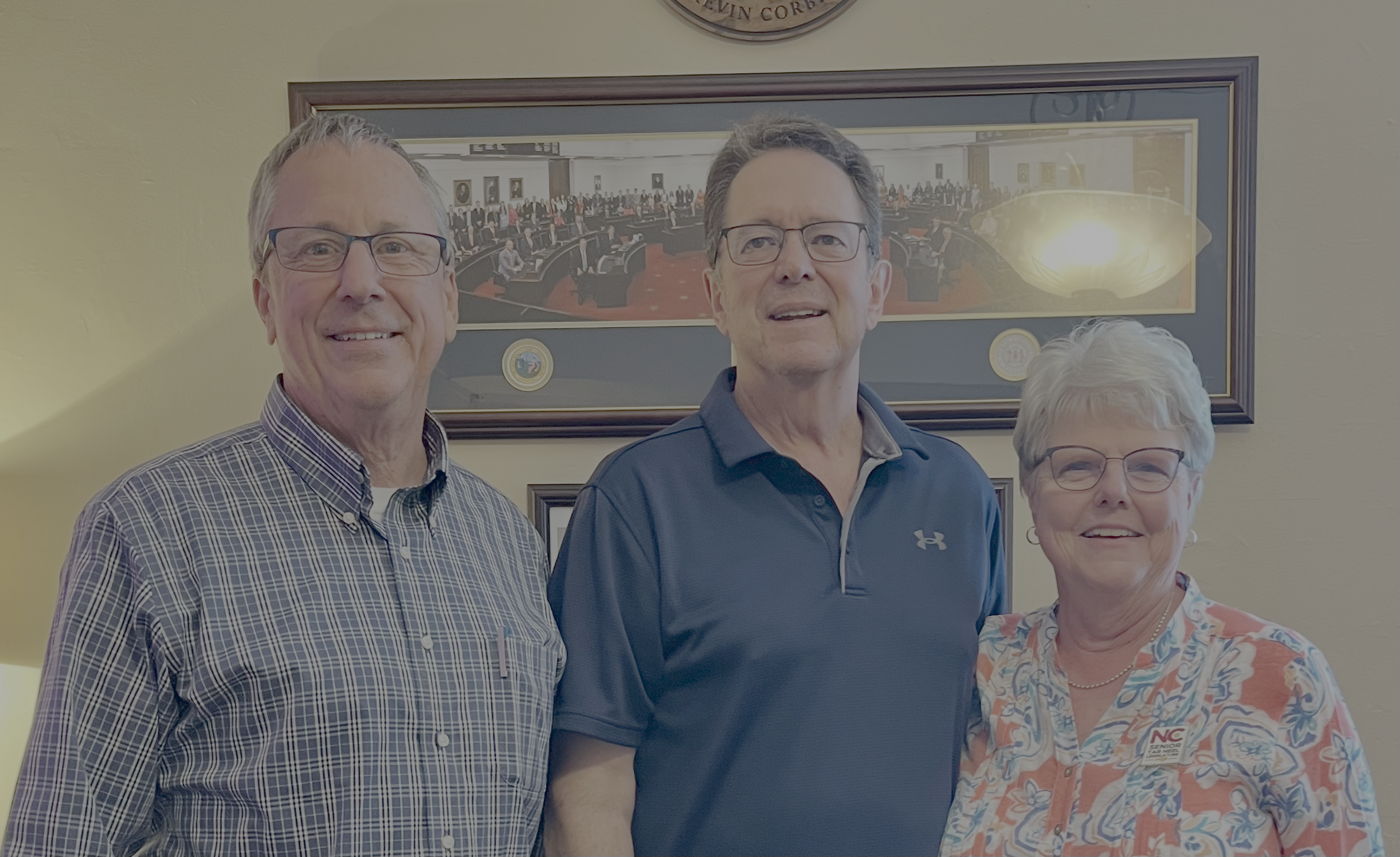 Three smiling people standing together in front of a framed painting of a legislative assembly room. The group includes two men and one woman, all wearing glasses.