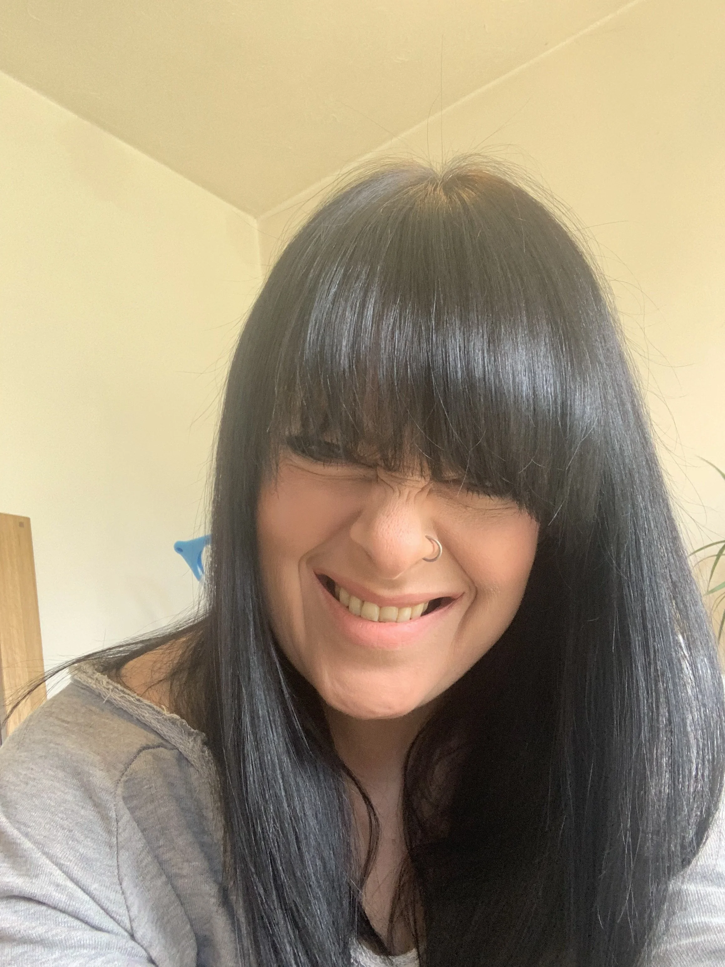 A smiling woman with straight black hair, wearing glasses and a nose ring, taking a selfie indoors.